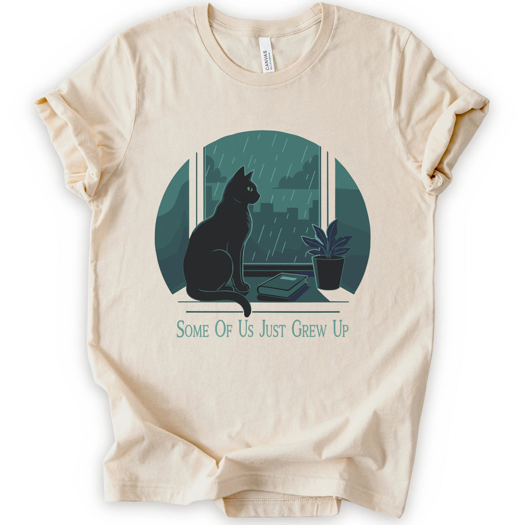 A natural short-sleeve crewneck t-shirt with a graphic print of a black cat sitting by a window in the rain, next to a small potted plant, with the text 'SOME OF US JUST GREW UP' printed below.