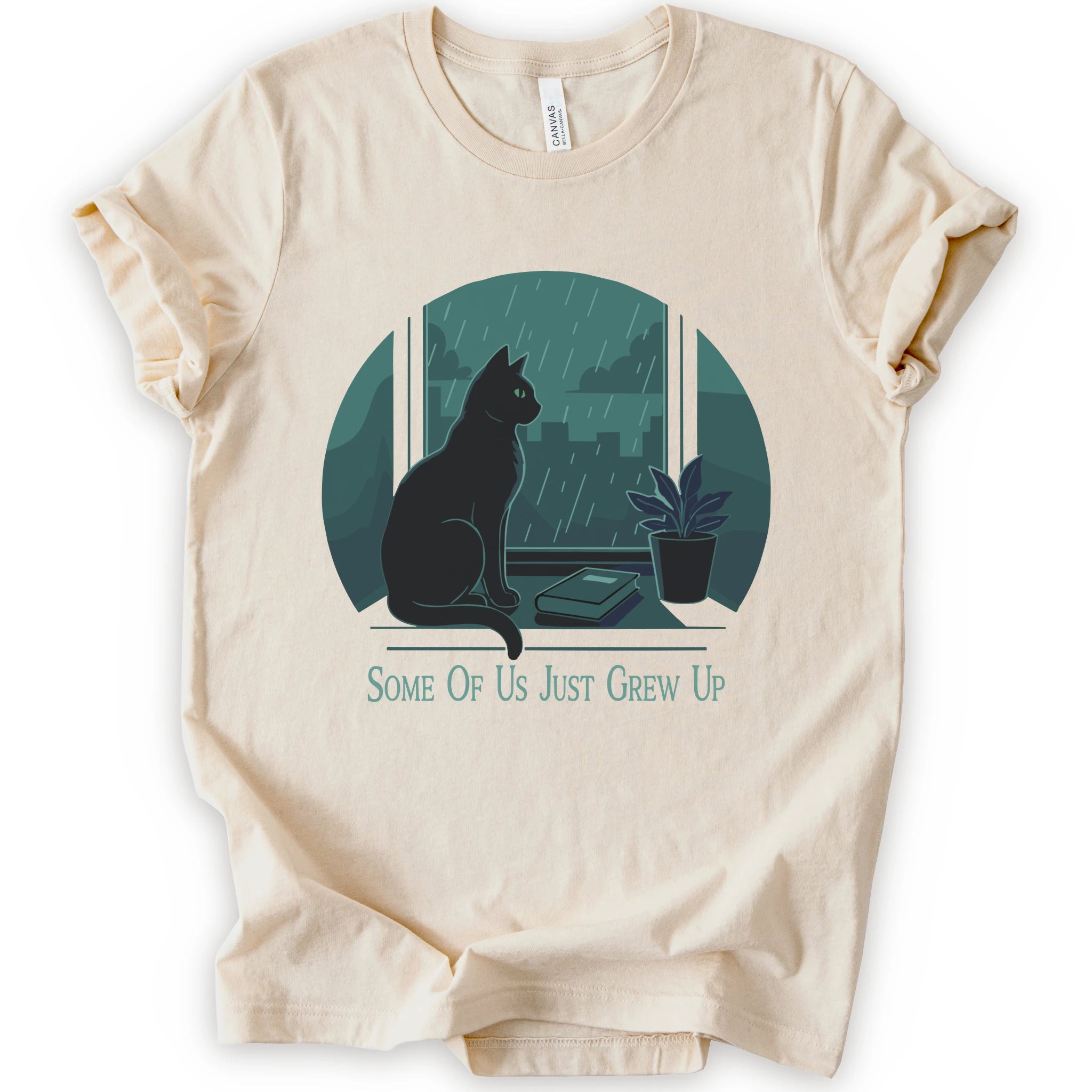 A natural short-sleeve crewneck t-shirt with a graphic print of a black cat sitting by a window in the rain, next to a small potted plant, with the text 'SOME OF US JUST GREW UP' printed below.