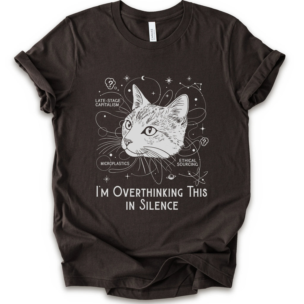 A brown short-sleeve crewneck t-shirt with a white graphic print of a cat's head surrounded by celestial and philosophical doodles, with the text 'I'M OVERTHINKING THIS IN SILENCE'.