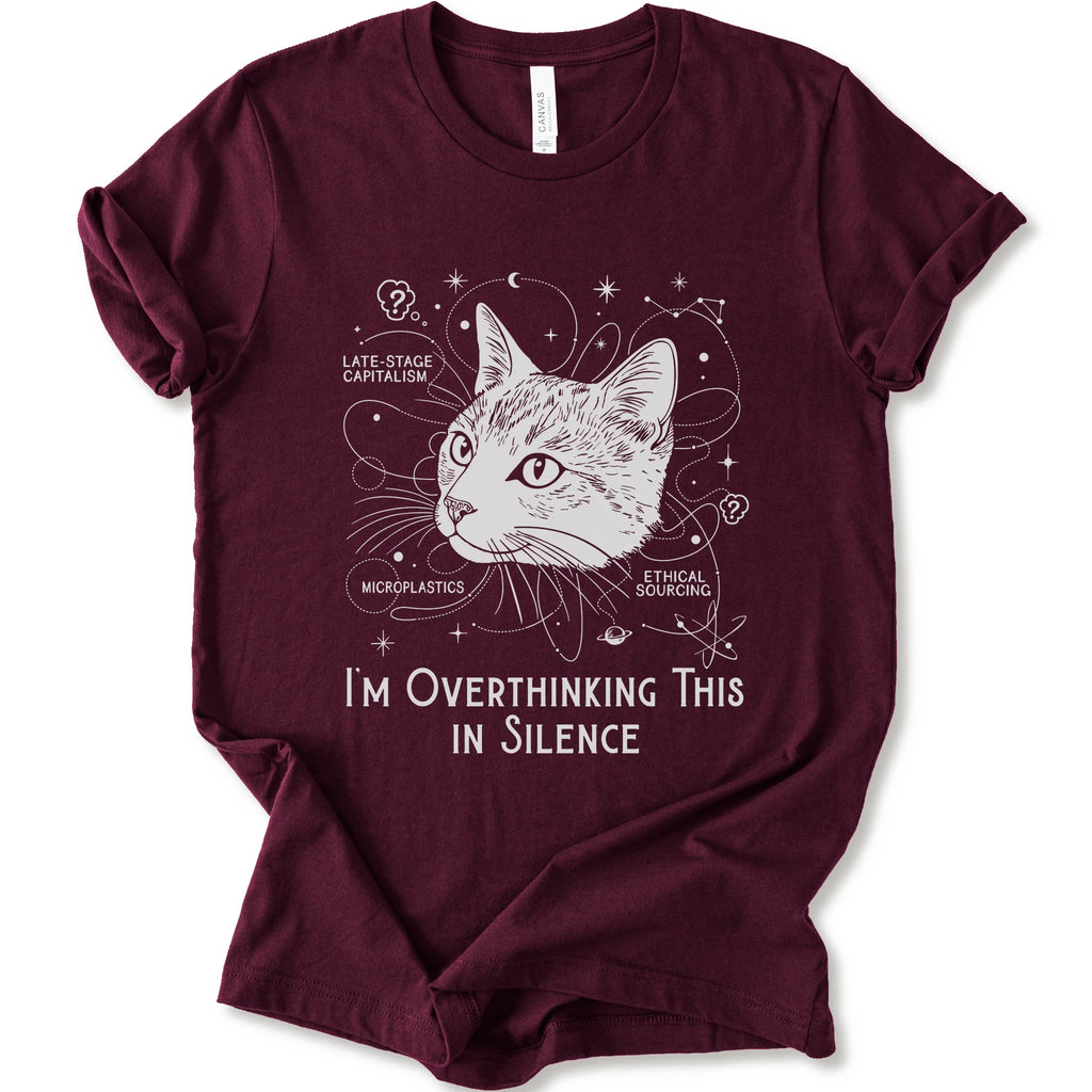 A maroon short-sleeve crewneck t-shirt with a white graphic print of a cat's head surrounded by celestial and philosophical doodles, with the text 'I'M OVERTHINKING THIS IN SILENCE'.