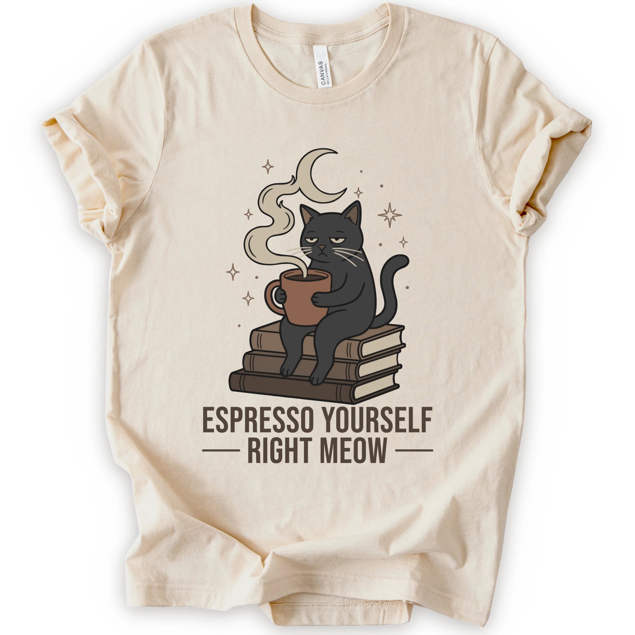 A natural short-sleeve crewneck t-shirt featuring a graphic of a black cat sitting on books and holding an espresso cup, with a crescent moon and stars above. The text 'ESPRESSO YOURSELF RIGHT MEOW' is printed below in brown.