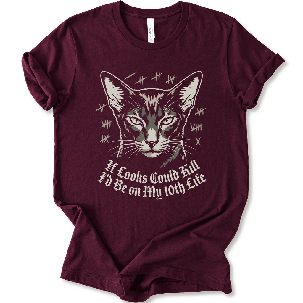 A maroon short-sleeve crewneck t-shirt featuring a graphic print of a cat's face with tally marks and the text 'It Looks Could Fill I'd Be on My 10th Life' in a white gothic font.