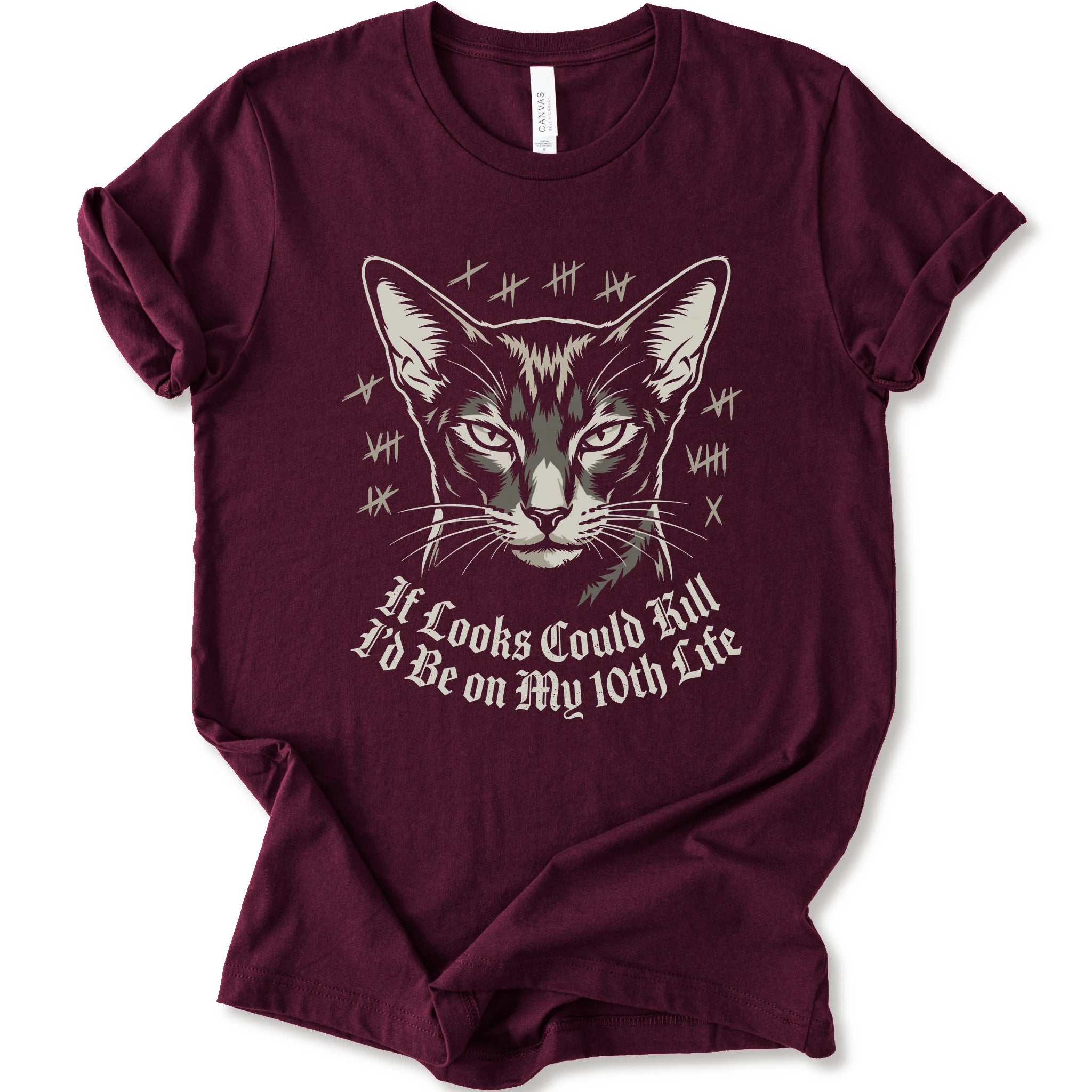 A maroon short-sleeve crewneck t-shirt featuring a graphic print of a cat's face with tally marks and the text 'It Looks Could Fill I'd Be on My 10th Life' in a white gothic font.