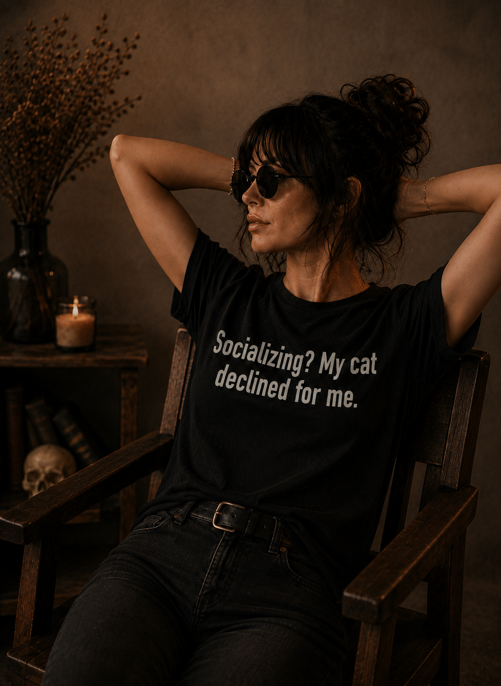 A woman wearing a black graphic t-shirt with the text 'Socializing? My cat declined for me.' sits in a wooden armchair in a dimly lit, rustic room.