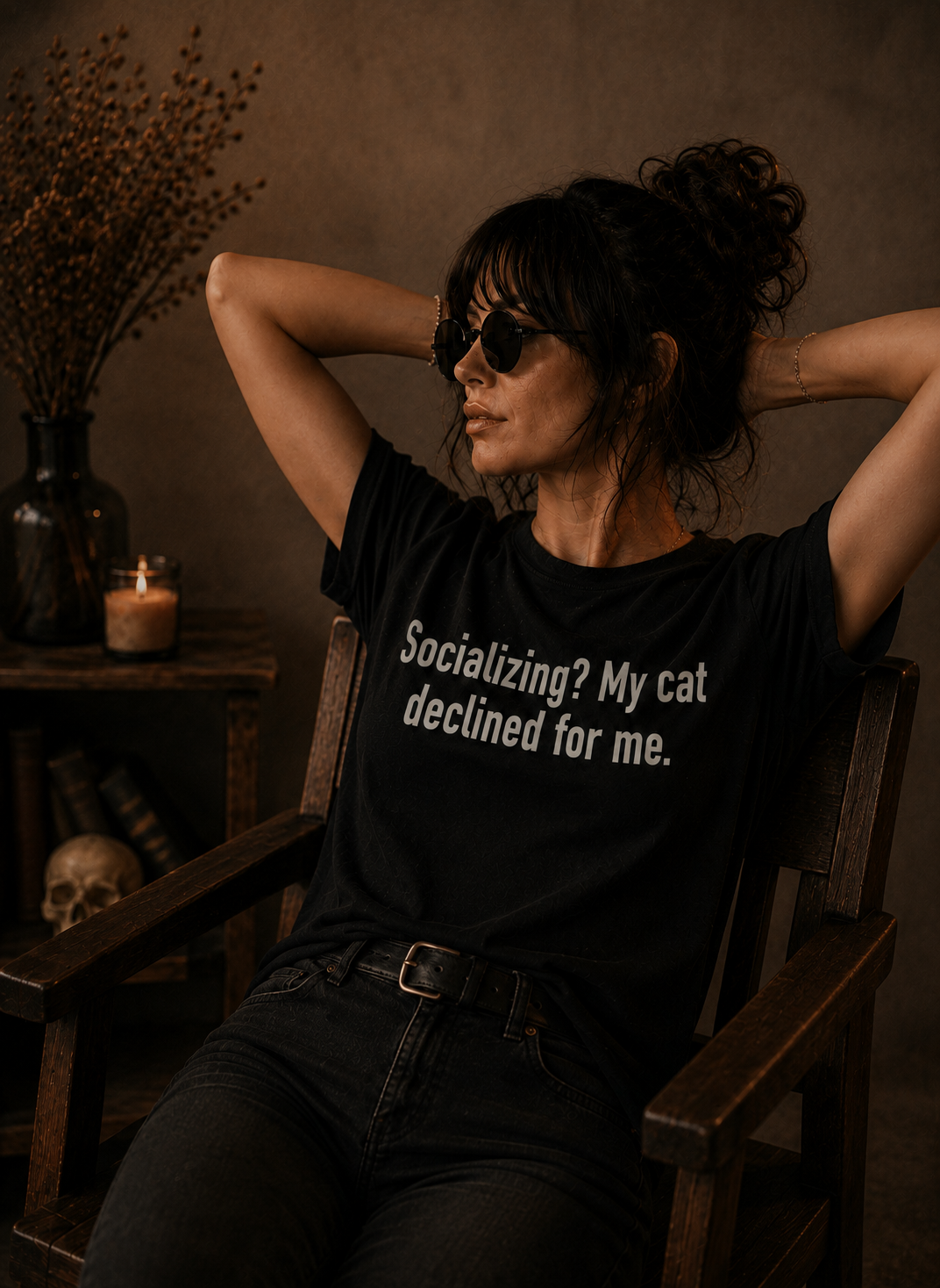 A woman wearing a black graphic t-shirt with the text 'Socializing? My cat declined for me.' sits in a wooden armchair in a dimly lit, rustic room.