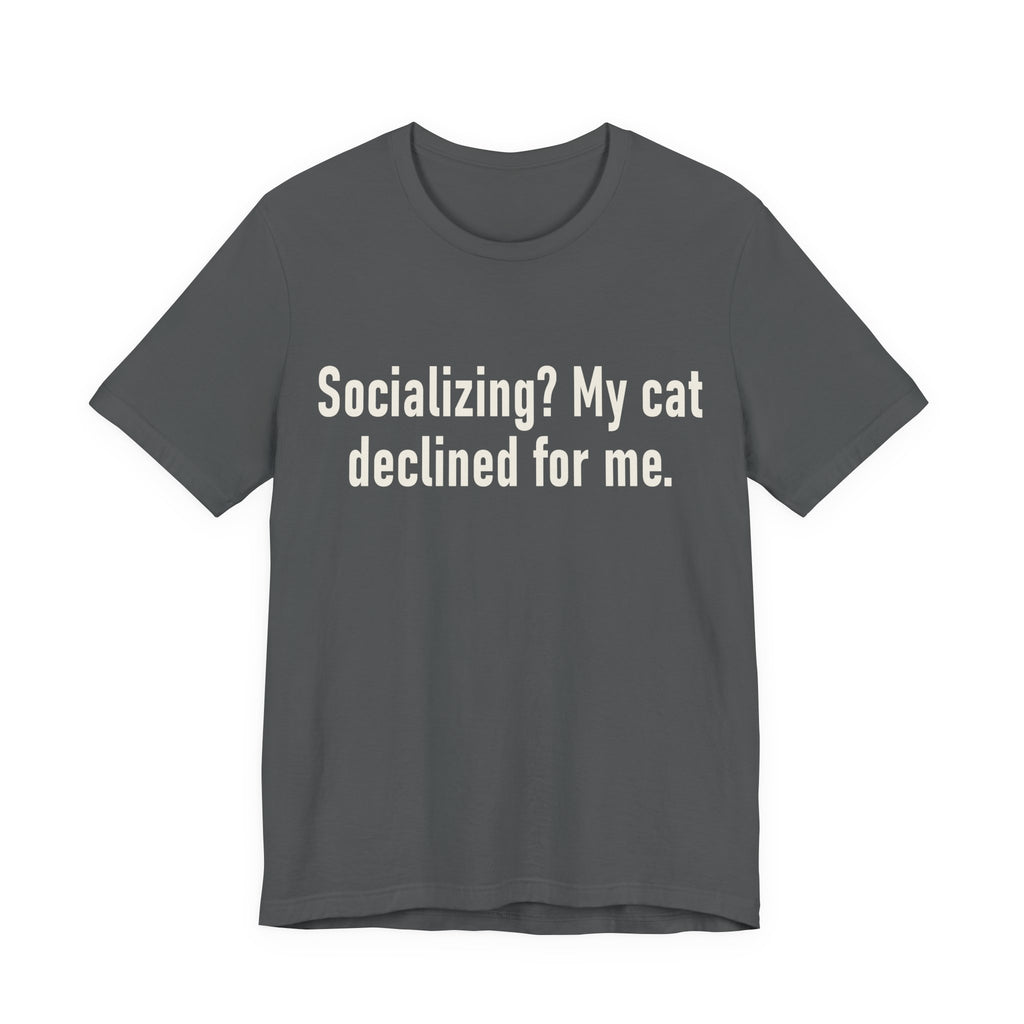 A asphalt short-sleeve crewneck t-shirt featuring a graphic with large cream-colored text reads “Socializing? My cat declined for me.” across two centered lines. The design is minimal, high-contrast and entirely typographic.