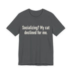 A asphalt short-sleeve crewneck t-shirt featuring a graphic with large cream-colored text reads “Socializing? My cat declined for me.” across two centered lines. The design is minimal, high-contrast and entirely typographic.