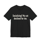 A black short-sleeve crewneck t-shirt featuring a graphic with large cream-colored text reads “Socializing? My cat declined for me.” across two centered lines. The design is minimal, high-contrast and entirely typographic.
