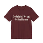 A maroon short-sleeve crewneck t-shirt featuring a graphic with large cream-colored text reads “Socializing? My cat declined for me.” across two centered lines. The design is minimal, high-contrast and entirely typographic.