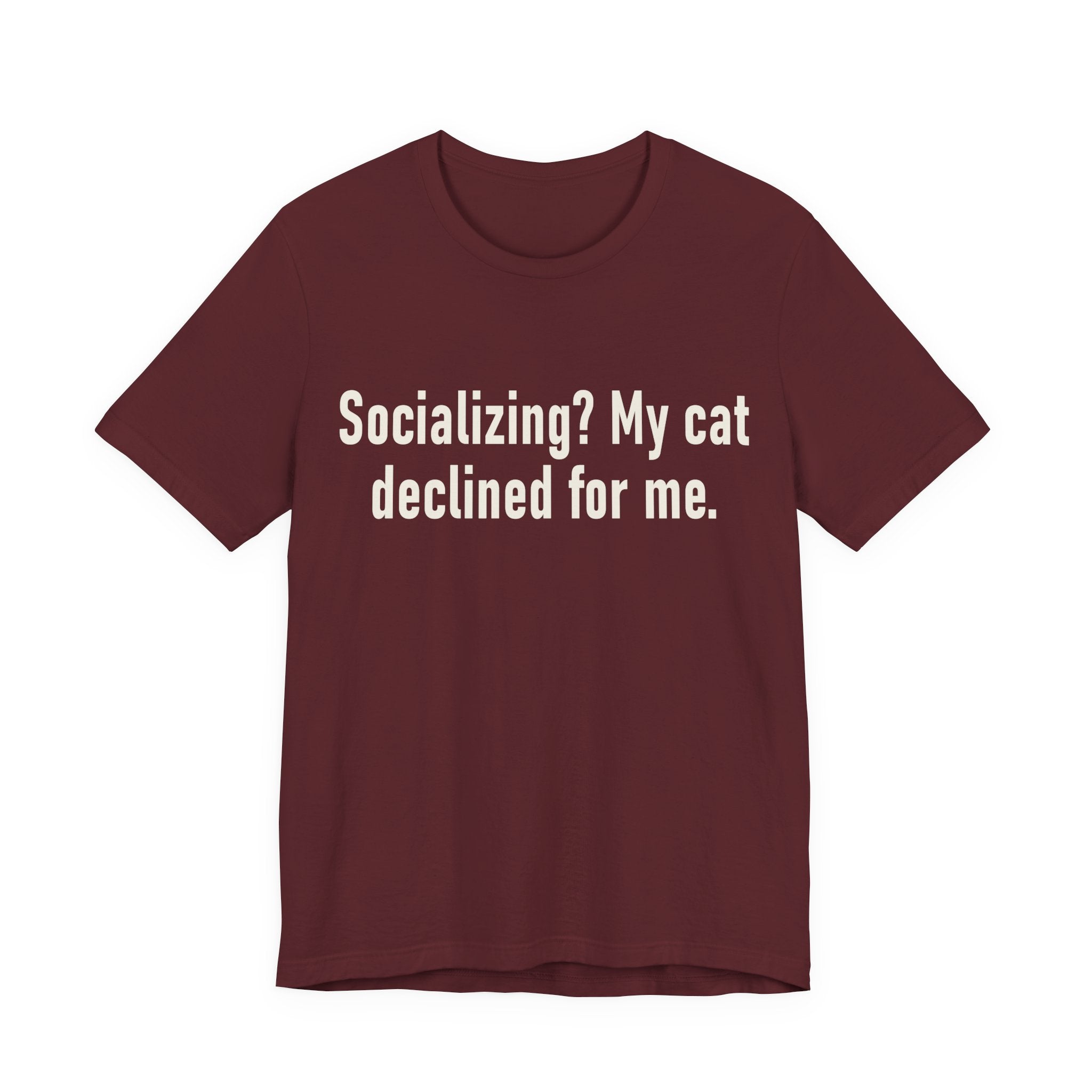 A maroon short-sleeve crewneck t-shirt featuring a graphic with large cream-colored text reads “Socializing? My cat declined for me.” across two centered lines. The design is minimal, high-contrast and entirely typographic.