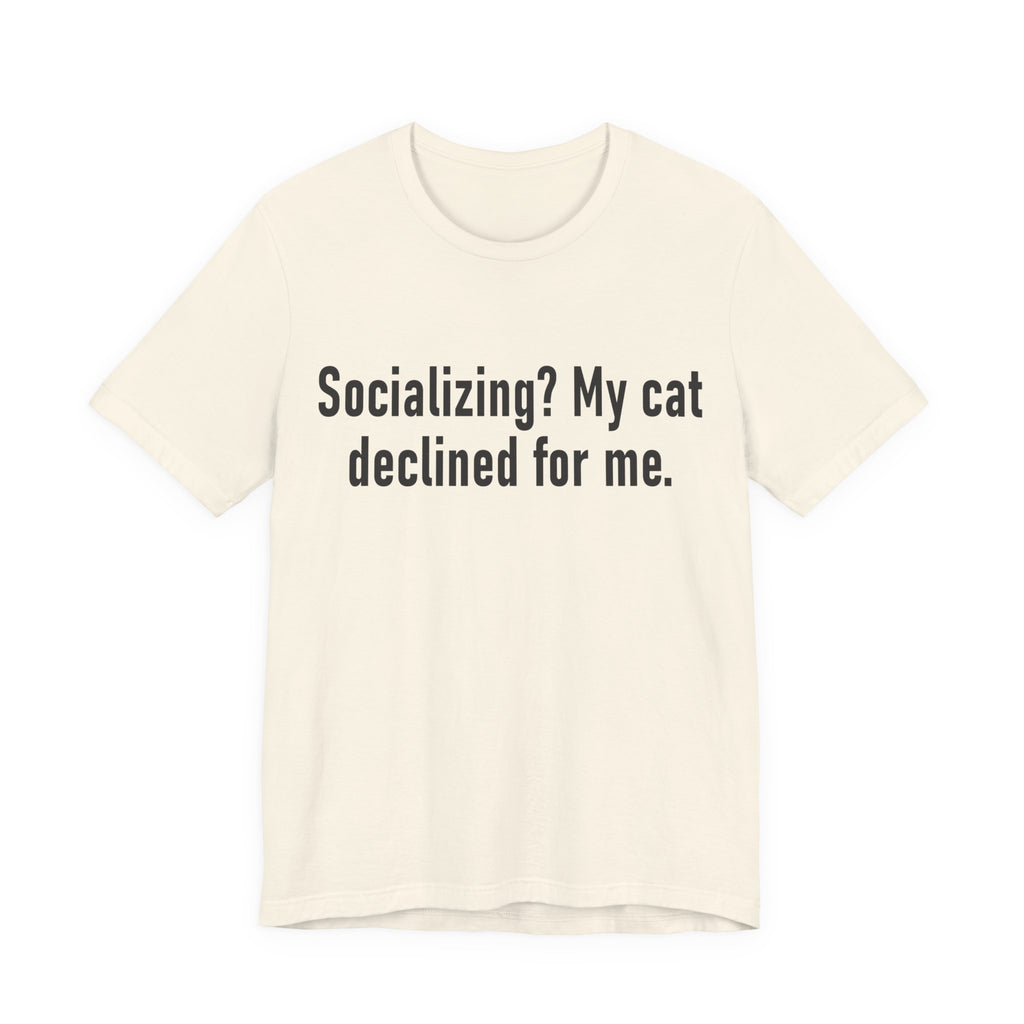 A natural short-sleeve crewneck t-shirt featuring a graphic with large black-colored text reads “Socializing? My cat declined for me.” across two centered lines. The design is minimal, high-contrast and entirely typographic.
