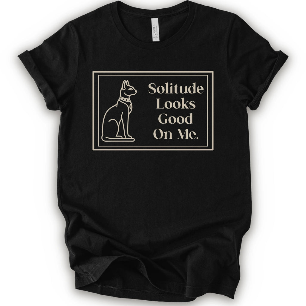 A black short-sleeve crewneck t-shirt with a cream-colored graphic on the front, features a thin rectangular border enclosing a line-art illustration of a cat on the left and the text 'Solitude Looks Good On Me.
