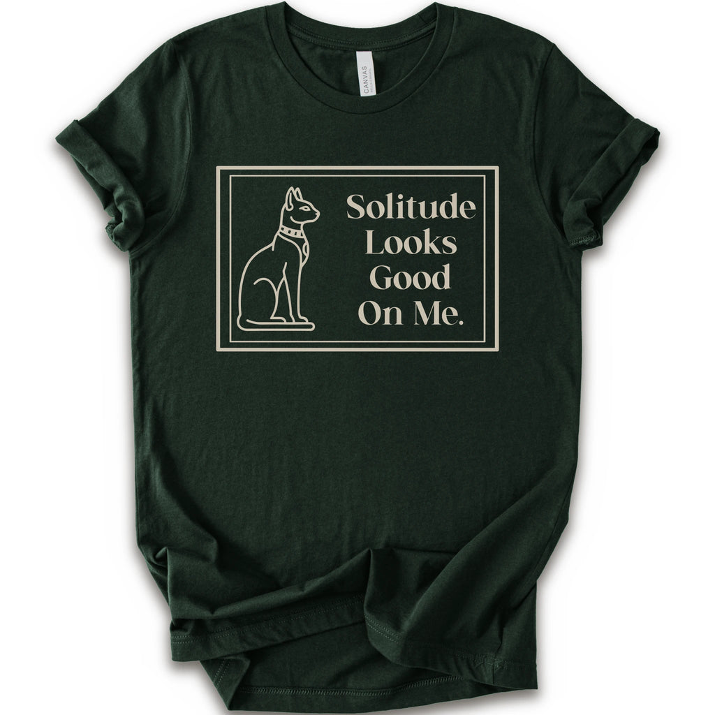 A forest short-sleeve crewneck t-shirt with a cream-colored graphic on the front, features a thin rectangular border enclosing a line-art illustration of a cat on the left and the text 'Solitude Looks Good On Me.
