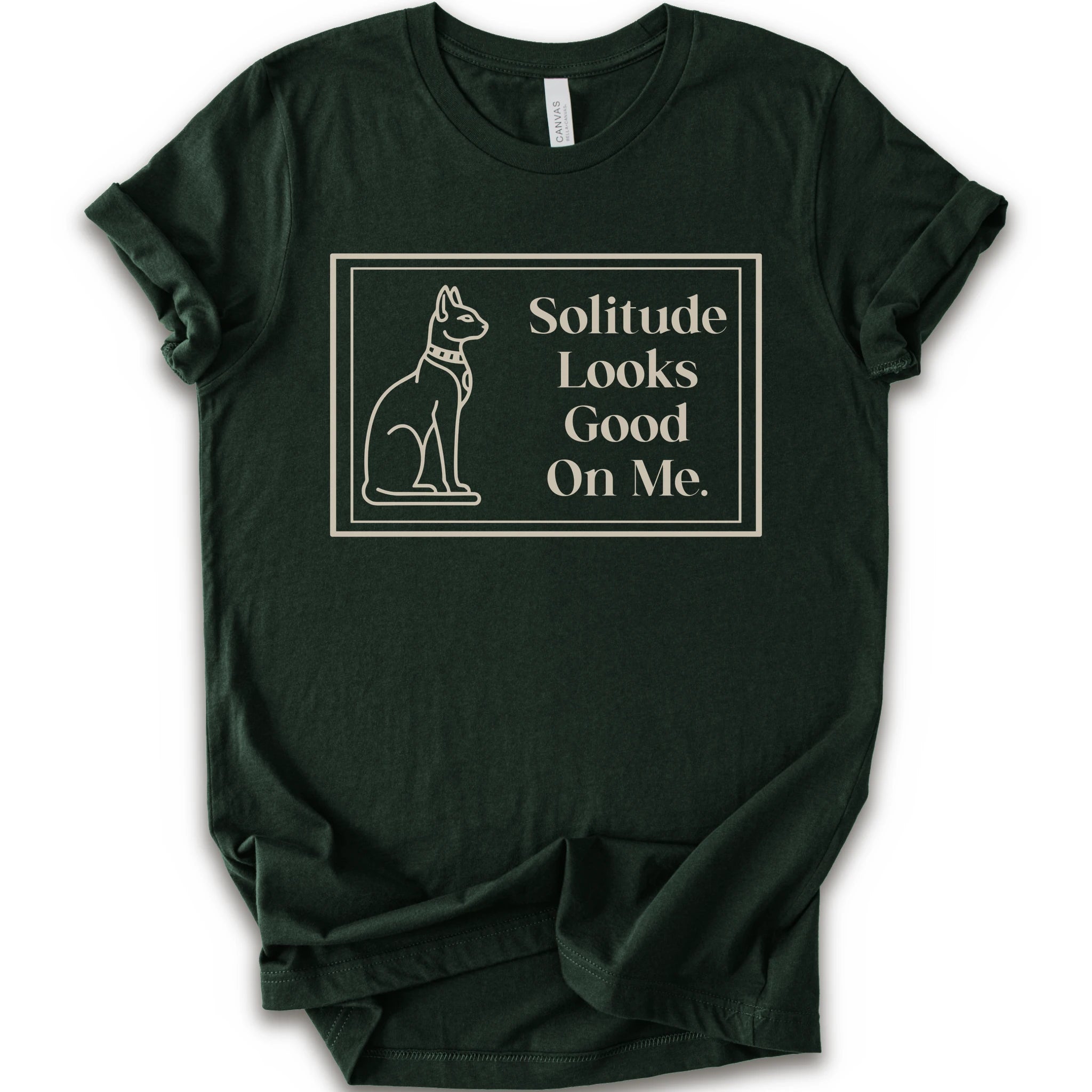 A forest short-sleeve crewneck t-shirt with a cream-colored graphic on the front, features a thin rectangular border enclosing a line-art illustration of a cat on the left and the text 'Solitude Looks Good On Me.