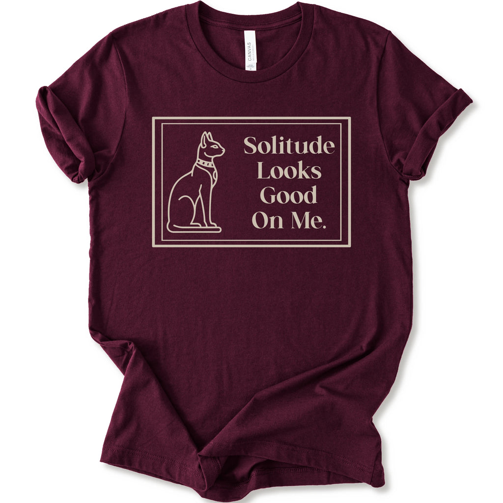 A maroon short-sleeve crewneck t-shirt with a cream-colored graphic on the front, features a thin rectangular border enclosing a line-art illustration of a cat on the left and the text 'Solitude Looks Good On Me.