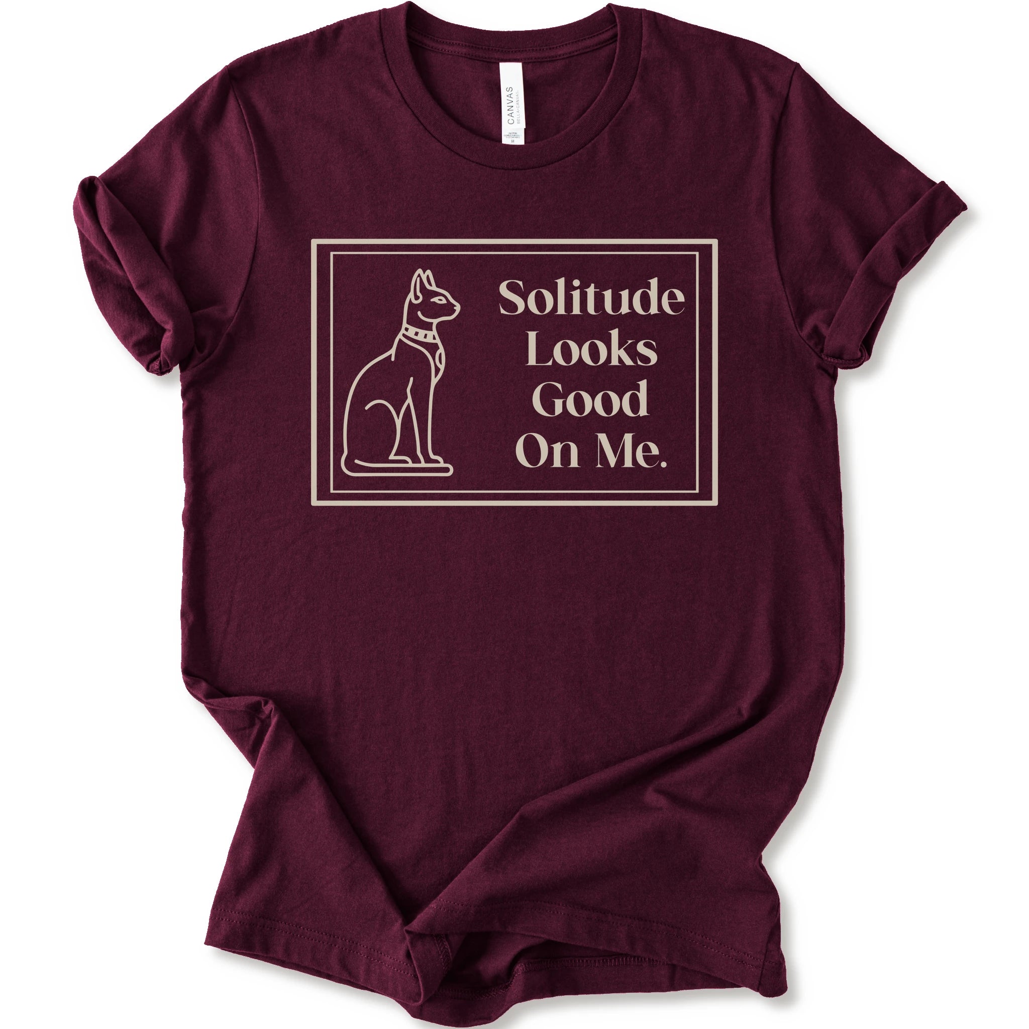 A maroon short-sleeve crewneck t-shirt with a cream-colored graphic on the front, features a thin rectangular border enclosing a line-art illustration of a cat on the left and the text 'Solitude Looks Good On Me.