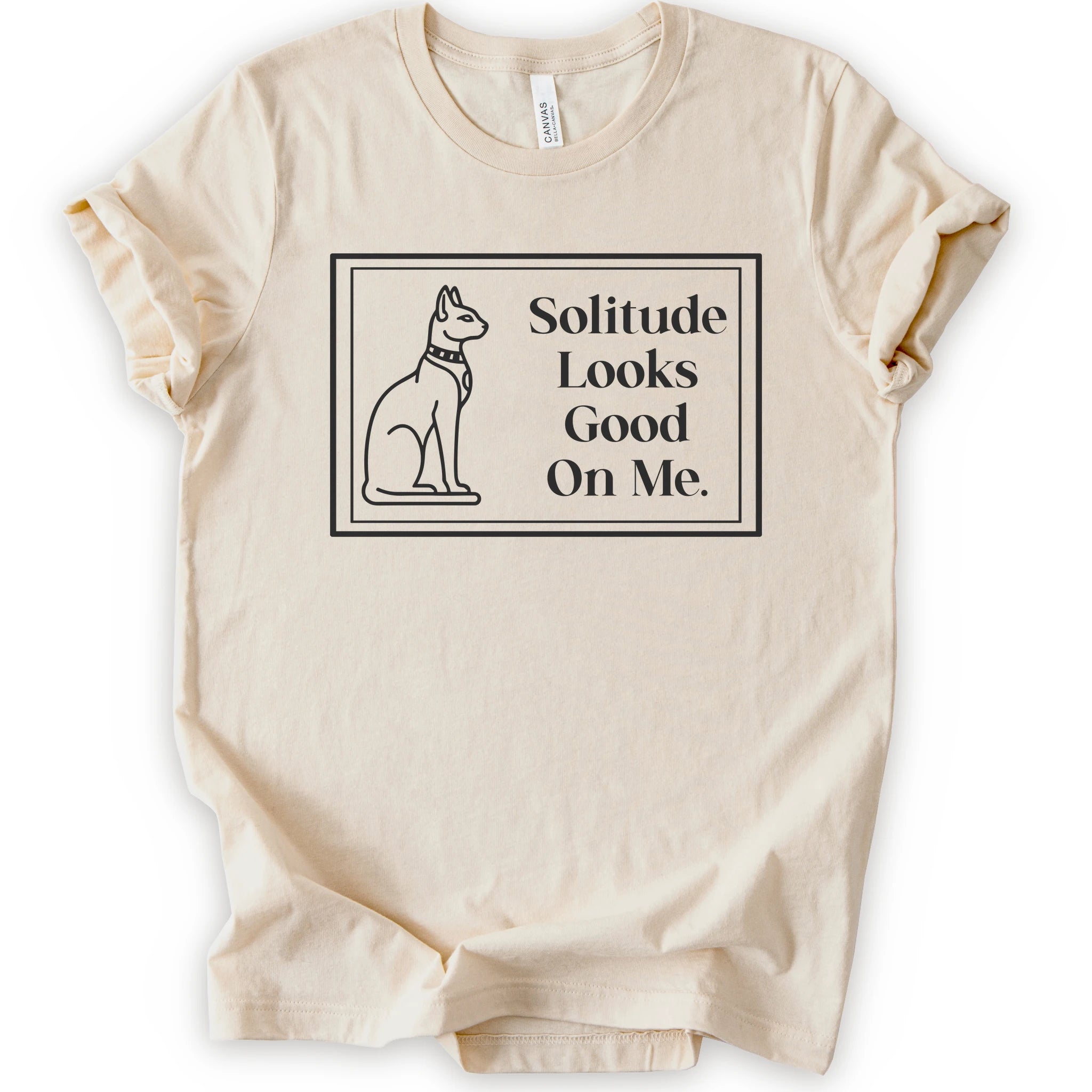 A natural short-sleeve crewneck t-shirt with a black-colored graphic on the front, features a thin rectangular border enclosing a line-art illustration of a cat on the left and the text 'Solitude Looks Good On Me.