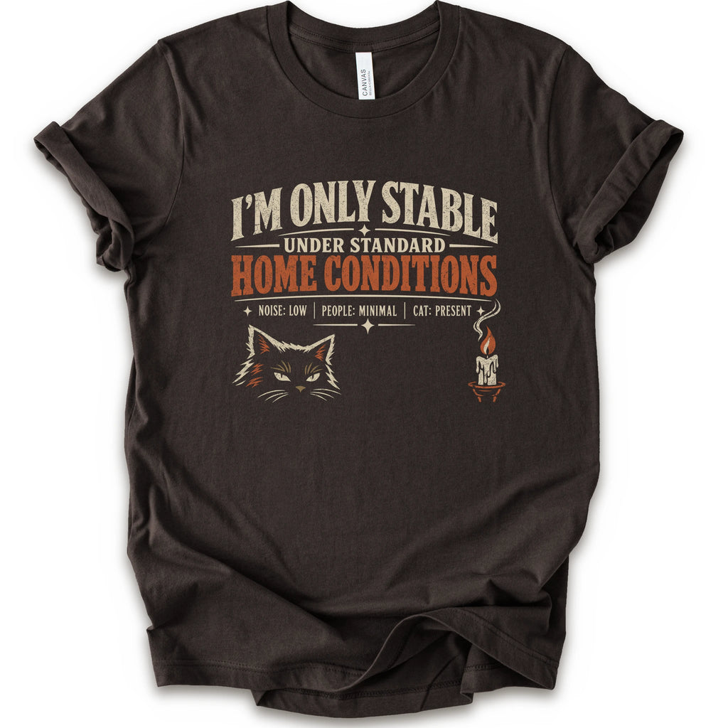 A brown short-sleeve graphic t-shirt features a humorous cat-themed print that reads 'I'M ONLY STABLE UNDER STANDARD HOME CONDITIONS' in cream and orange lettering, accompanied by a graphic of a cat's face and a small candle with a cat inside.