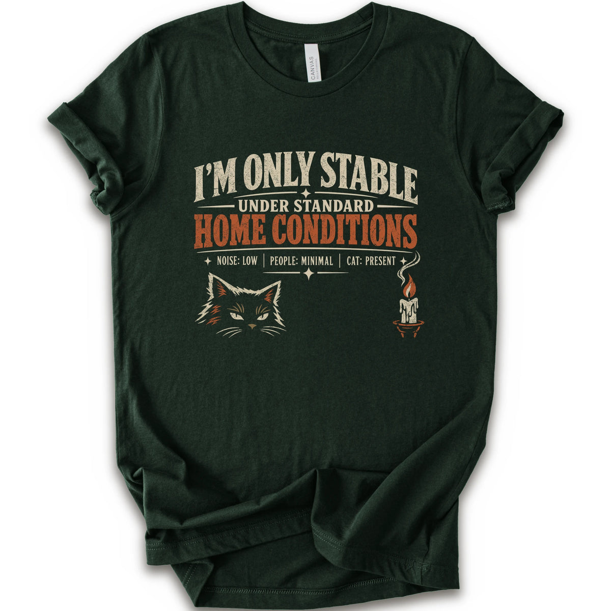 A forest short-sleeve graphic t-shirt features a humorous cat-themed print that reads 'I'M ONLY STABLE UNDER STANDARD HOME CONDITIONS' in cream and orange lettering, accompanied by a graphic of a cat's face and a small candle with a cat inside.