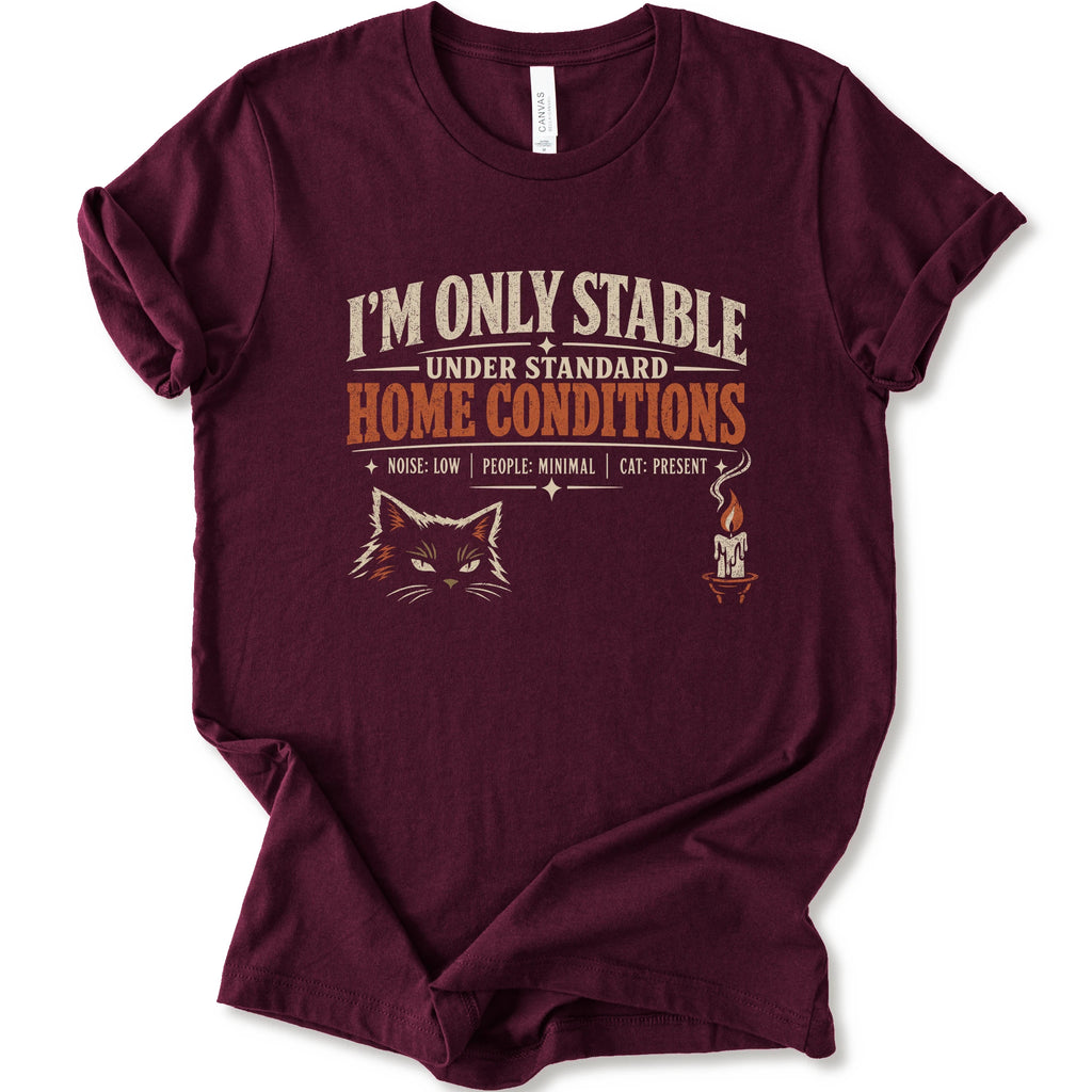A maroon short-sleeve graphic t-shirt features a humorous cat-themed print that reads 'I'M ONLY STABLE UNDER STANDARD HOME CONDITIONS' in cream and orange lettering, accompanied by a graphic of a cat's face and a small candle with a cat inside.