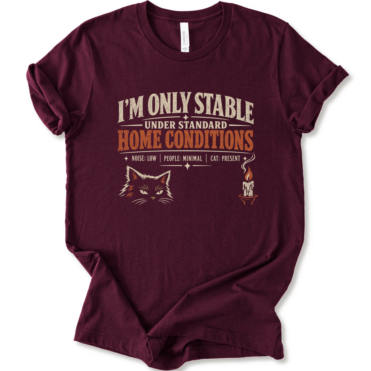 A maroon short-sleeve graphic t-shirt features a humorous cat-themed print that reads 'I'M ONLY STABLE UNDER STANDARD HOME CONDITIONS' in cream and orange lettering, accompanied by a graphic of a cat's face and a small candle with a cat inside.