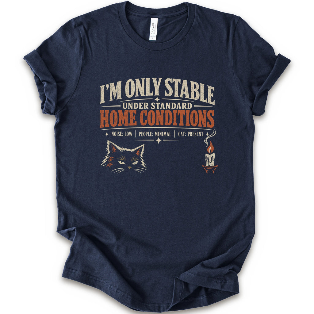 A navy short-sleeve graphic t-shirt features a humorous cat-themed print that reads 'I'M ONLY STABLE UNDER STANDARD HOME CONDITIONS' in cream and orange lettering, accompanied by a graphic of a cat's face and a small candle with a cat inside.