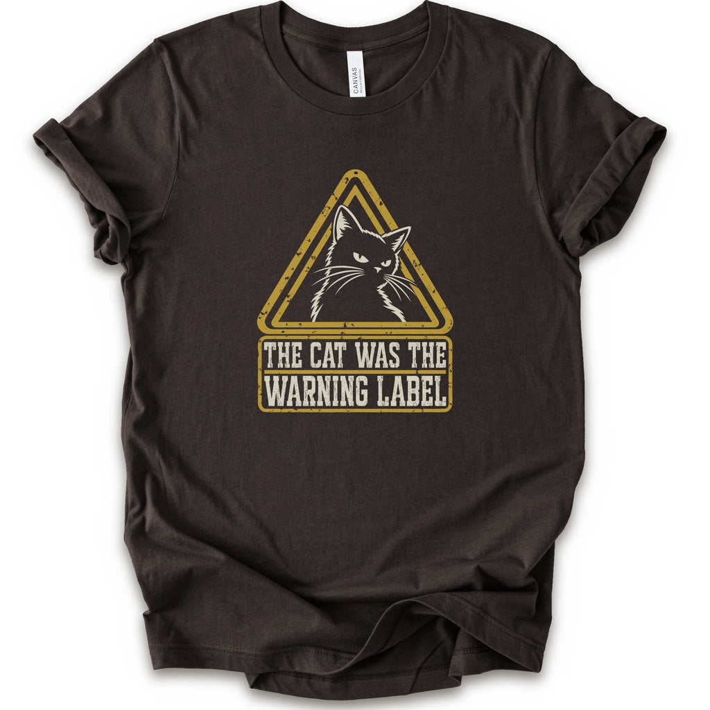 A brown short-sleeve crewneck t-shirt with a distressed yellow and white graphic on the front showing a cat inside a warning triangle and the text 'THE CAT WAS THE WARNING LABEL'.