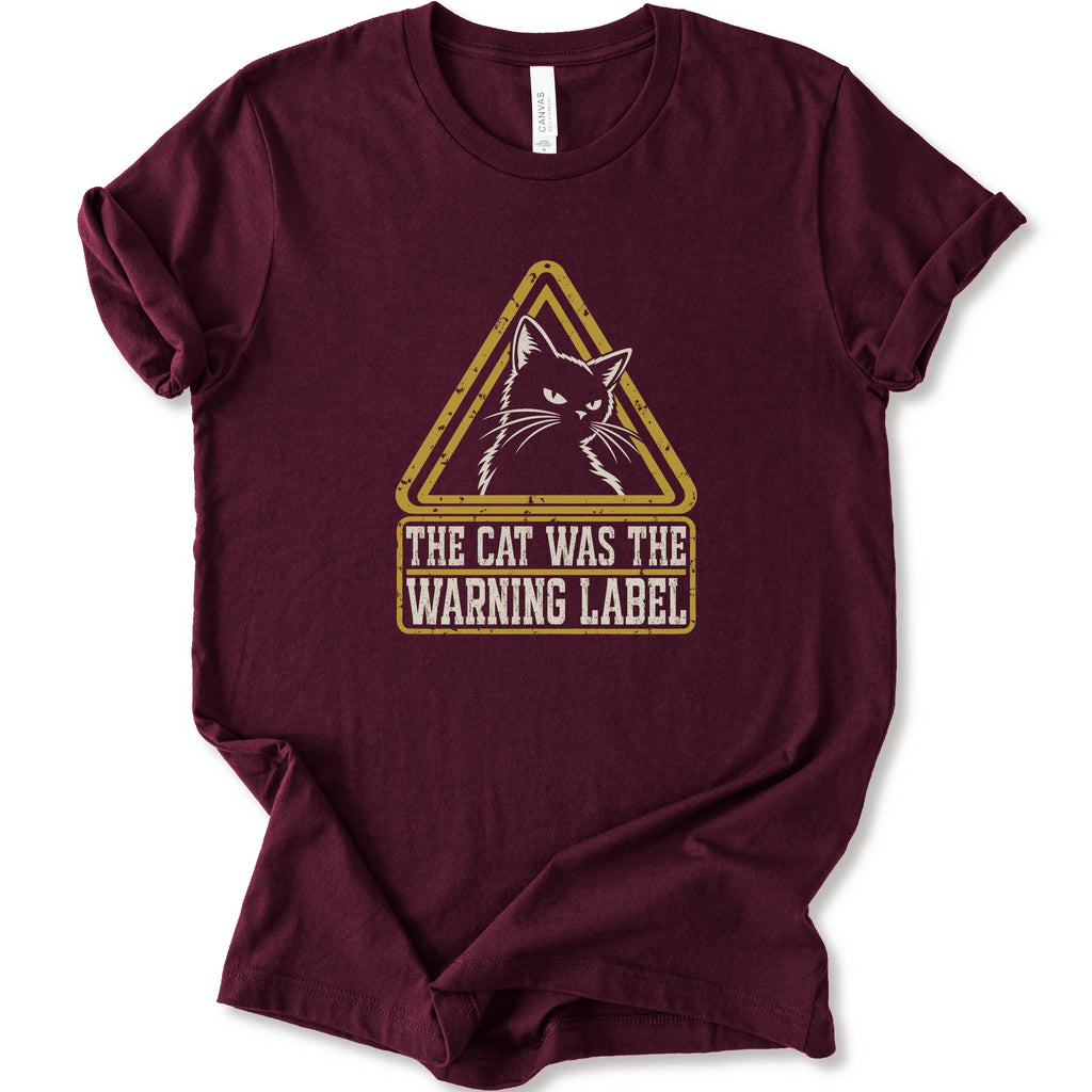 A maroon short-sleeve crewneck t-shirt with a distressed yellow and white graphic on the front showing a cat inside a warning triangle and the text 'THE CAT WAS THE WARNING LABEL'.