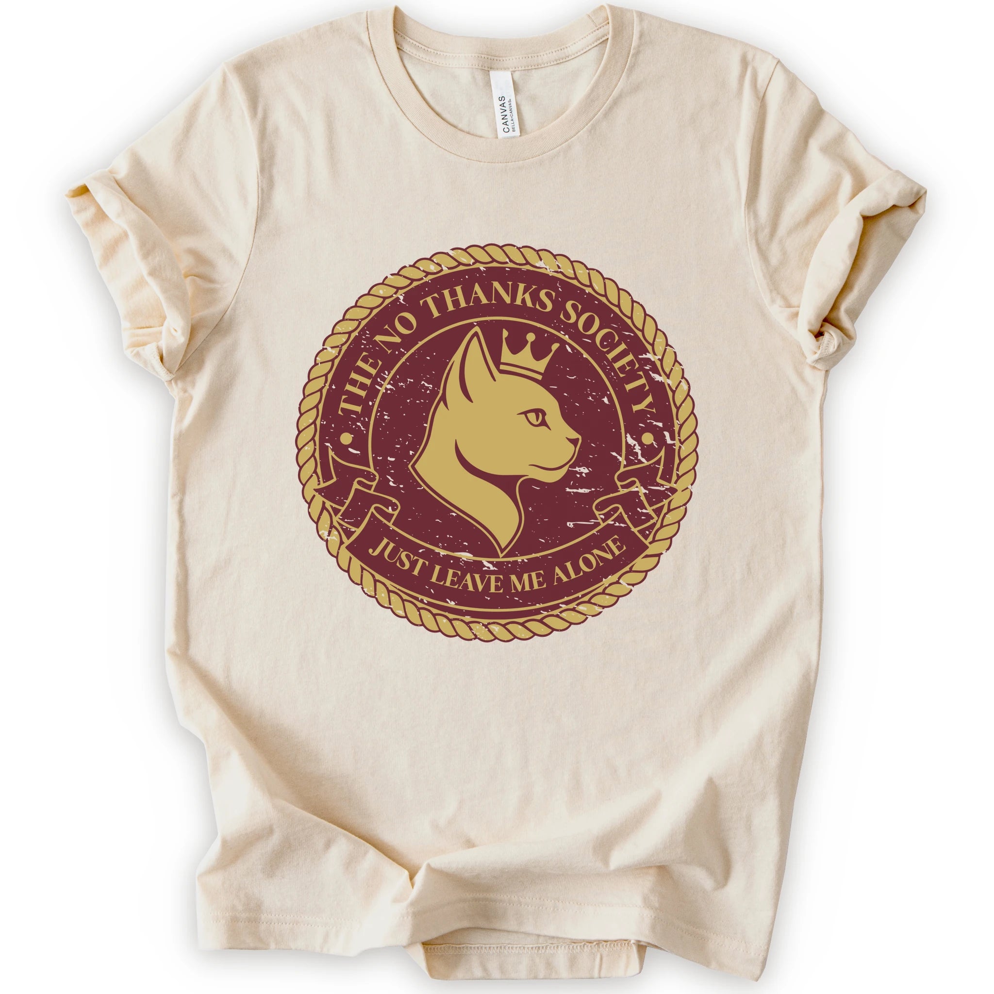 A natural short-sleeve t-shirt with a circular graphic print on the front. The graphic features a golden-yellow cat profile wearing a crown, surrounded by a rope border and the text 'THE NO THANKS SOCIETY' and 'JUST LEAVE ME ALONE' in a maroon background.