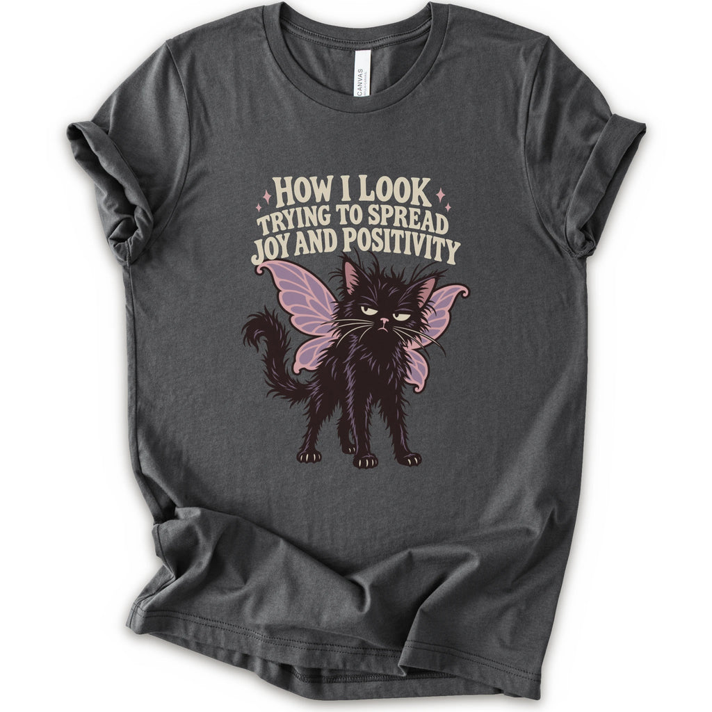 An asphalt short-sleeve crewneck t-shirt with a graphic of a black cat with pink-purple fairy wings and the text 'HOW I LOOK TRYING TO SPREAD JOY AND POSITIVITY' in a light beige font.