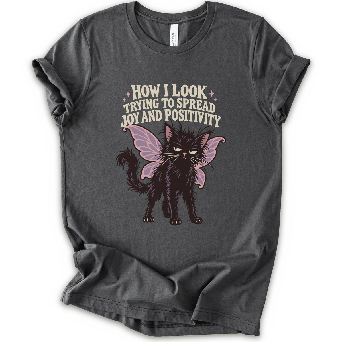 An asphalt short-sleeve crewneck t-shirt with a graphic of a black cat with pink-purple fairy wings and the text 'HOW I LOOK TRYING TO SPREAD JOY AND POSITIVITY' in a light beige font.