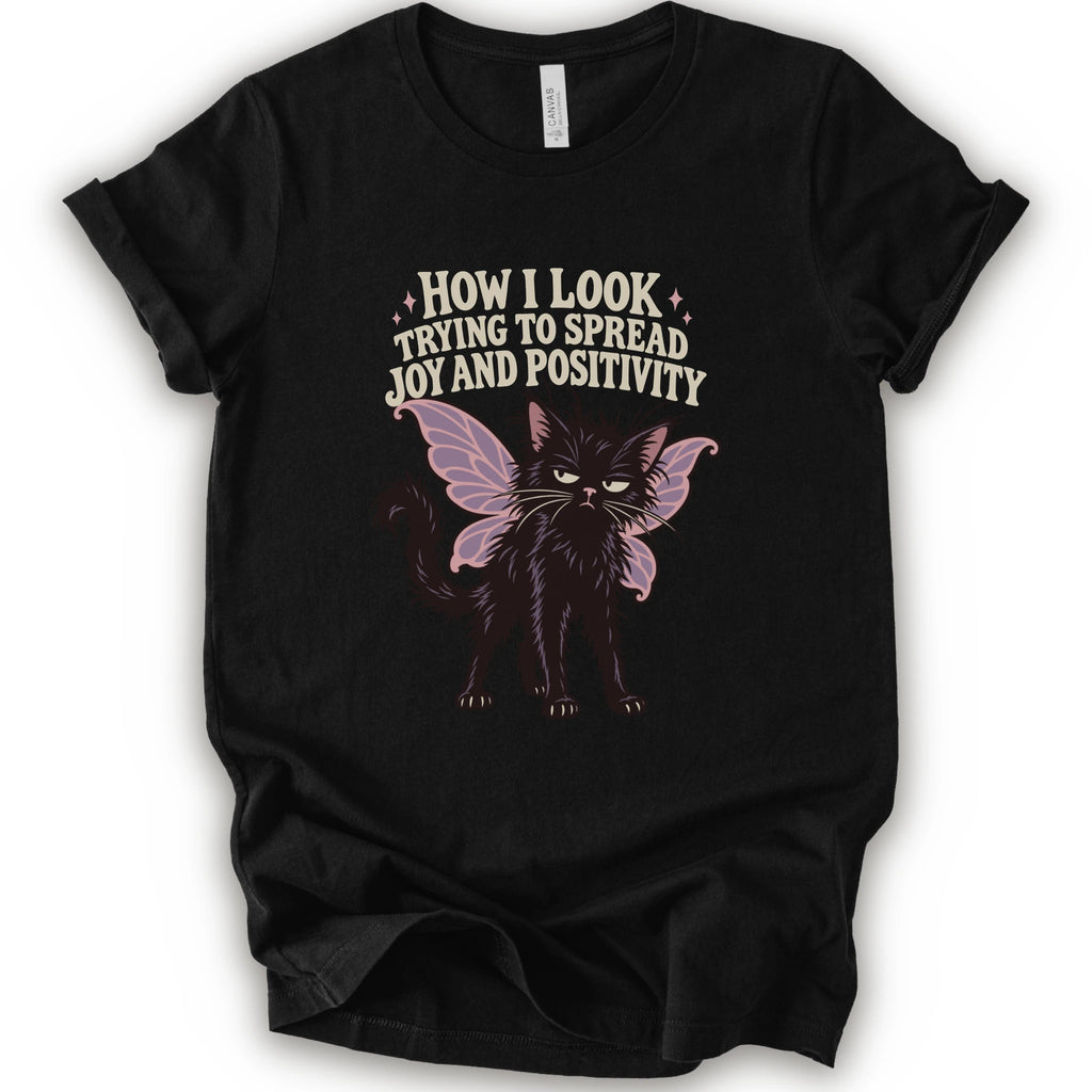 A black short-sleeve crewneck t-shirt with a graphic of a black cat with pink-purple fairy wings and the text 'HOW I LOOK TRYING TO SPREAD JOY AND POSITIVITY' in a light beige font.