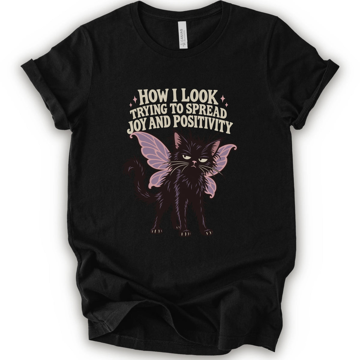 A black short-sleeve crewneck t-shirt with a graphic of a black cat with pink-purple fairy wings and the text 'HOW I LOOK TRYING TO SPREAD JOY AND POSITIVITY' in a light beige font.