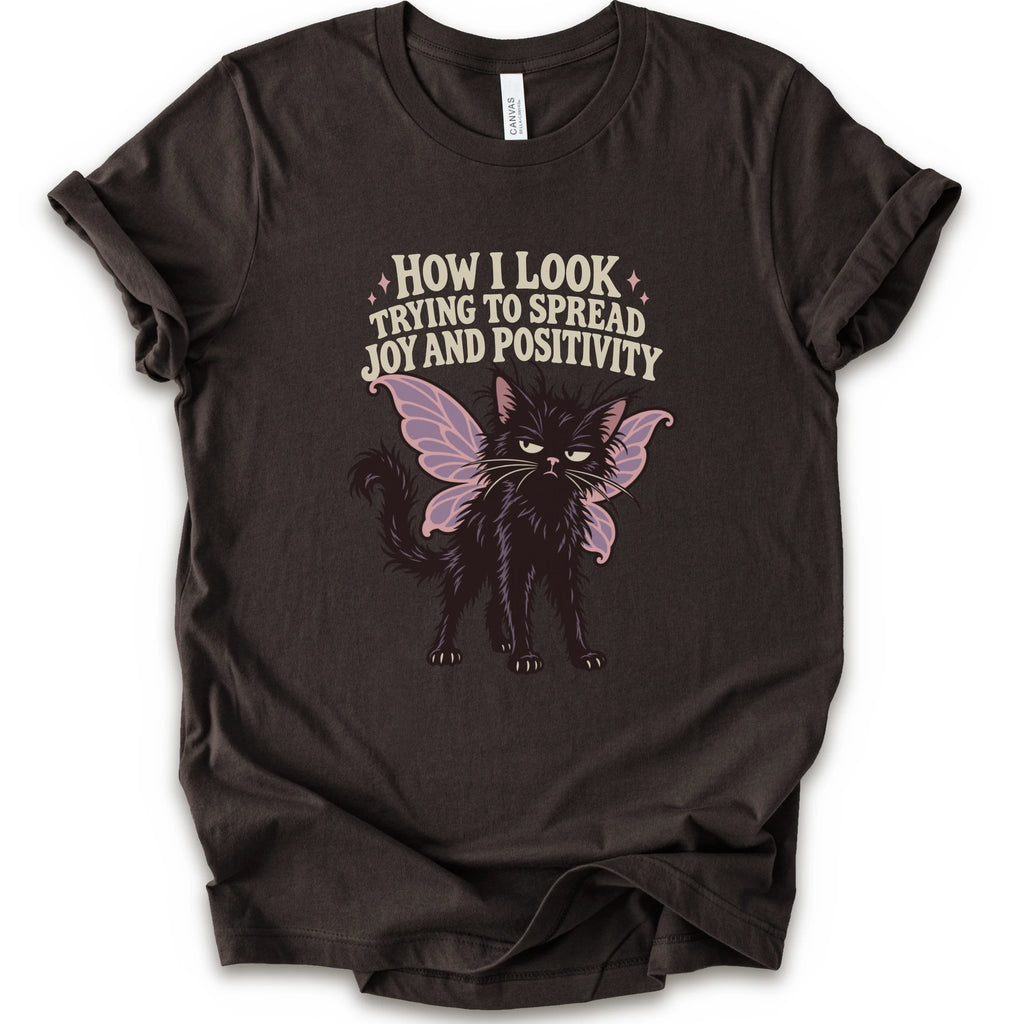 A brown short-sleeve crewneck t-shirt with a graphic of a black cat with pink-purple fairy wings and the text 'HOW I LOOK TRYING TO SPREAD JOY AND POSITIVITY' in a light beige font.