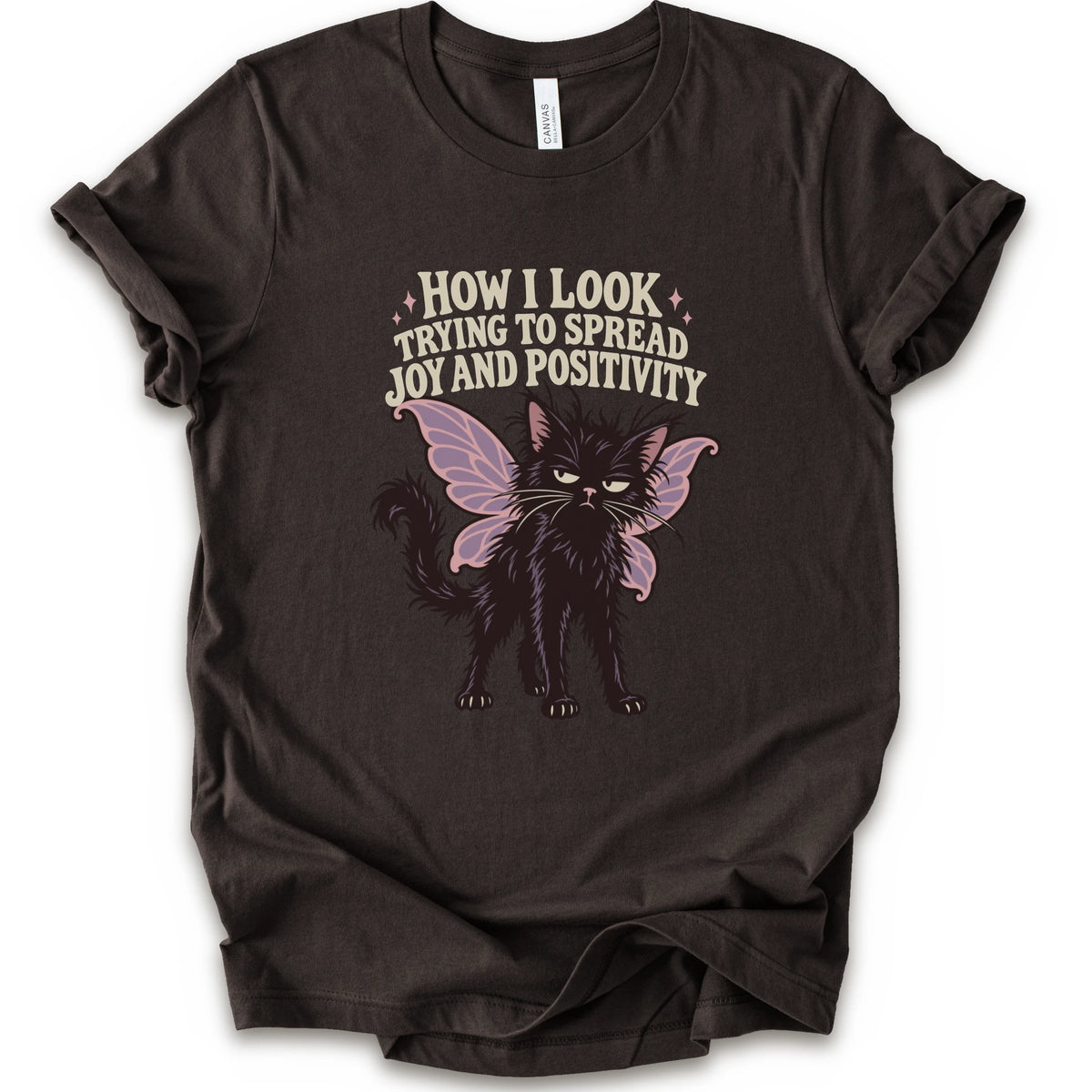 A brown short-sleeve crewneck t-shirt with a graphic of a black cat with pink-purple fairy wings and the text 'HOW I LOOK TRYING TO SPREAD JOY AND POSITIVITY' in a light beige font.