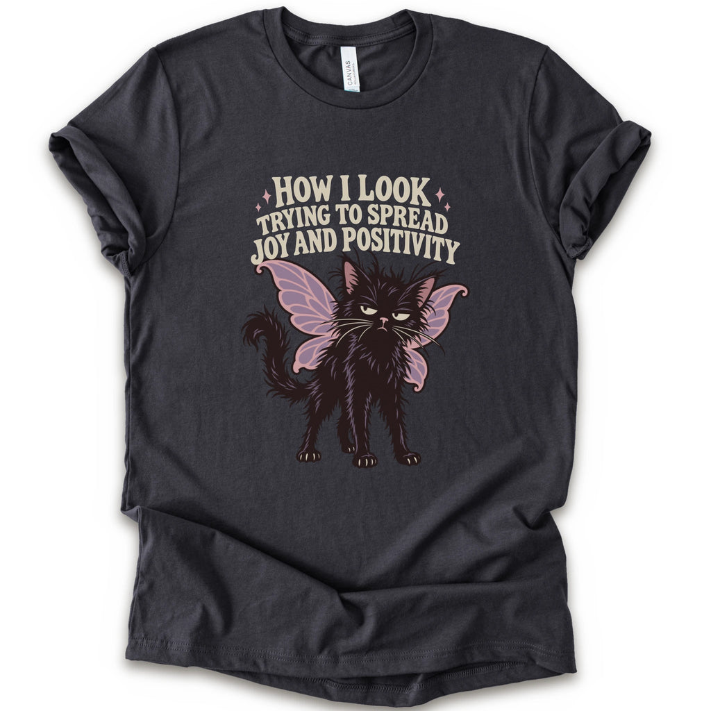 A dark grey short-sleeve crewneck t-shirt with a graphic of a black cat with pink-purple fairy wings and the text 'HOW I LOOK TRYING TO SPREAD JOY AND POSITIVITY' in a light beige font.