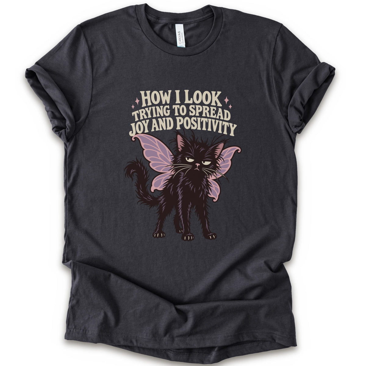 A dark grey short-sleeve crewneck t-shirt with a graphic of a black cat with pink-purple fairy wings and the text 'HOW I LOOK TRYING TO SPREAD JOY AND POSITIVITY' in a light beige font.