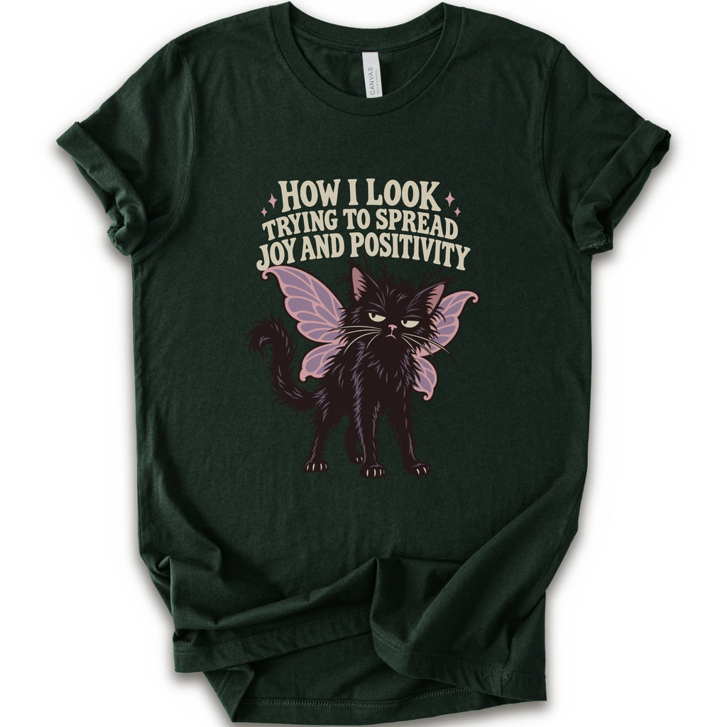 A forest short-sleeve crewneck t-shirt with a graphic of a black cat with pink-purple fairy wings and the text 'HOW I LOOK TRYING TO SPREAD JOY AND POSITIVITY' in a light beige font.