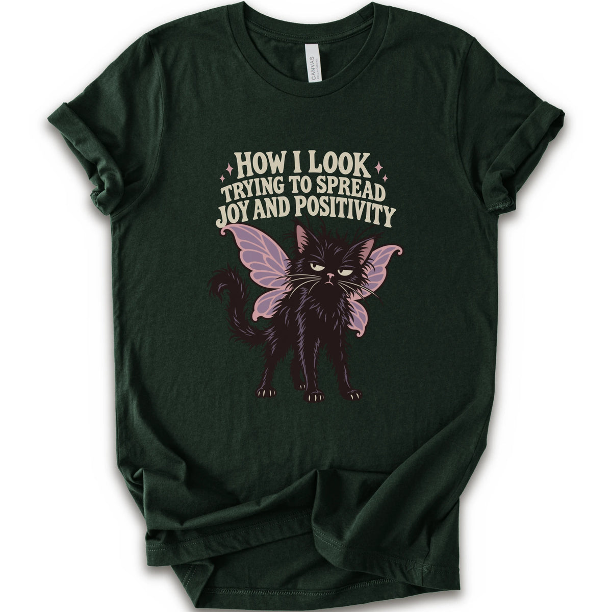 A forest short-sleeve crewneck t-shirt with a graphic of a black cat with pink-purple fairy wings and the text 'HOW I LOOK TRYING TO SPREAD JOY AND POSITIVITY' in a light beige font.