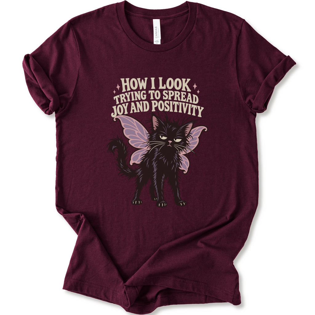 A maroon short-sleeve crewneck t-shirt with a graphic of a black cat with pink-purple fairy wings and the text 'HOW I LOOK TRYING TO SPREAD JOY AND POSITIVITY' in a light beige font.