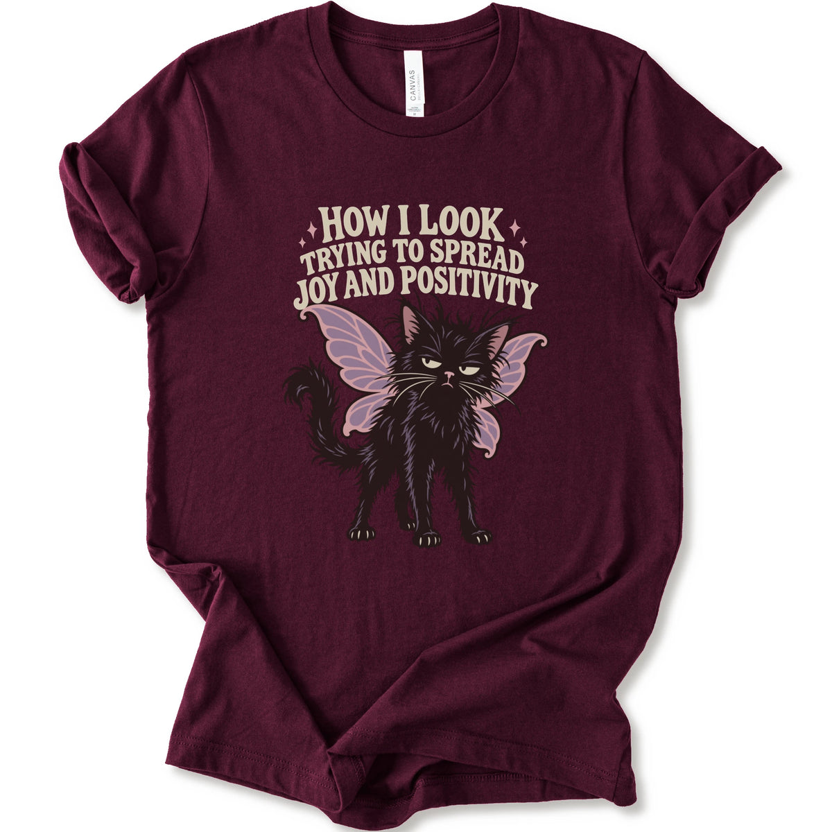 A maroon short-sleeve crewneck t-shirt with a graphic of a black cat with pink-purple fairy wings and the text 'HOW I LOOK TRYING TO SPREAD JOY AND POSITIVITY' in a light beige font.