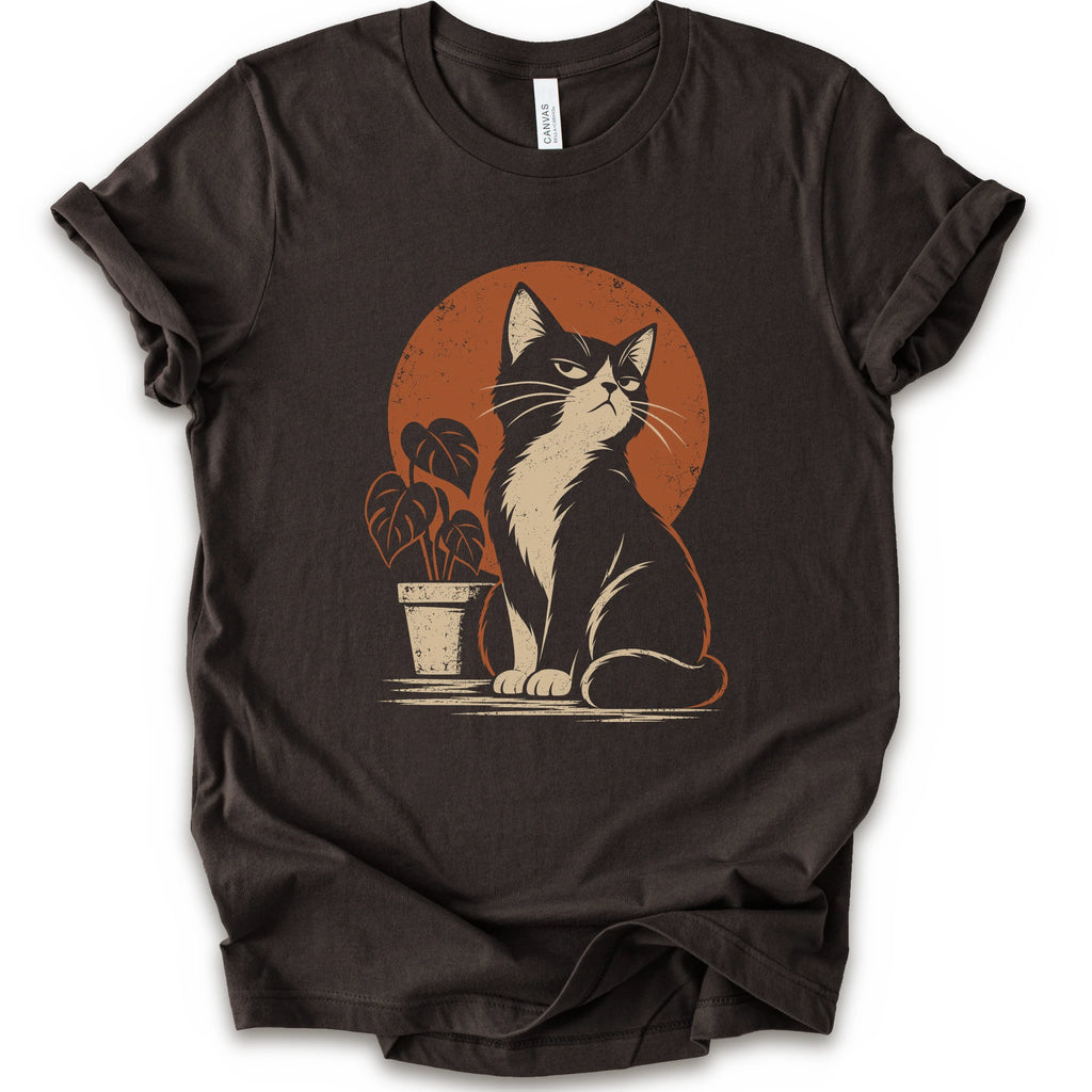 A brown short-sleeve crewneck t-shirt with a retro-style graphic of a black and white cat sitting in front of an orange circular sun, next to a potted plant.