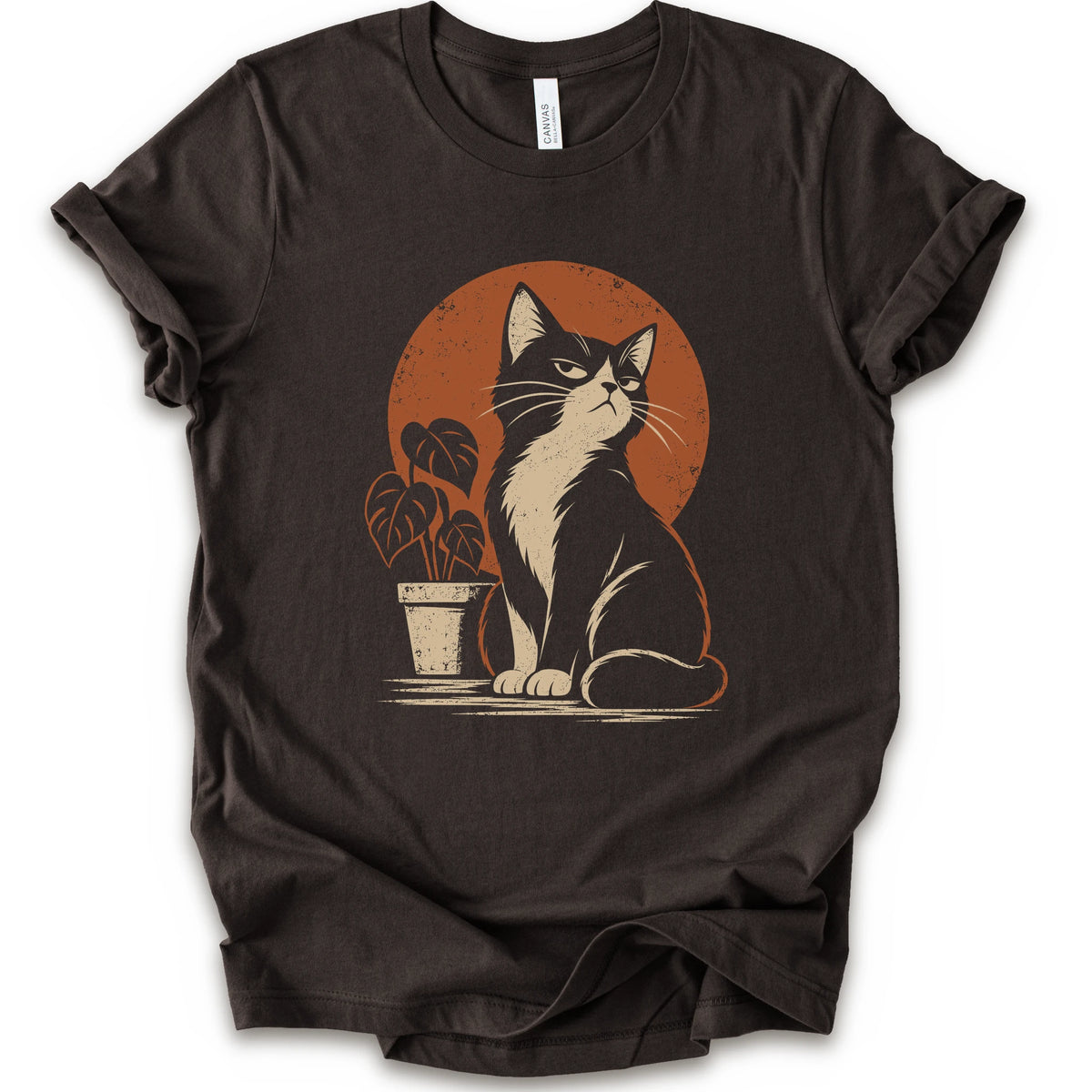 A brown short-sleeve crewneck t-shirt with a retro-style graphic of a black and white cat sitting in front of an orange circular sun, next to a potted plant.