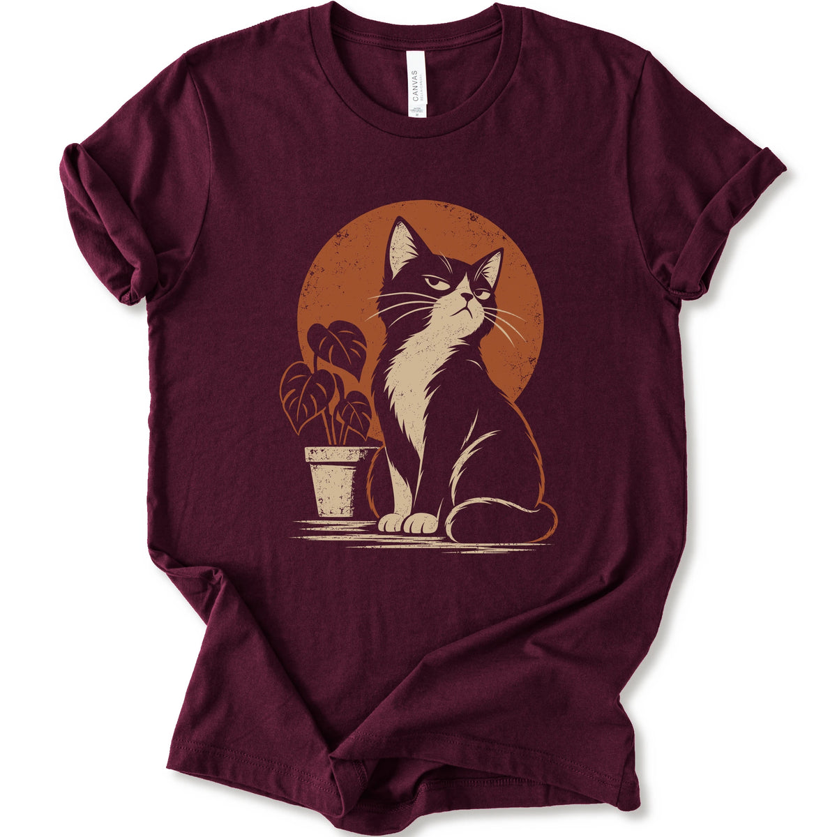 A maroon short-sleeve crewneck t-shirt with a retro-style graphic of a black and white cat sitting in front of an orange circular sun, next to a potted plant.