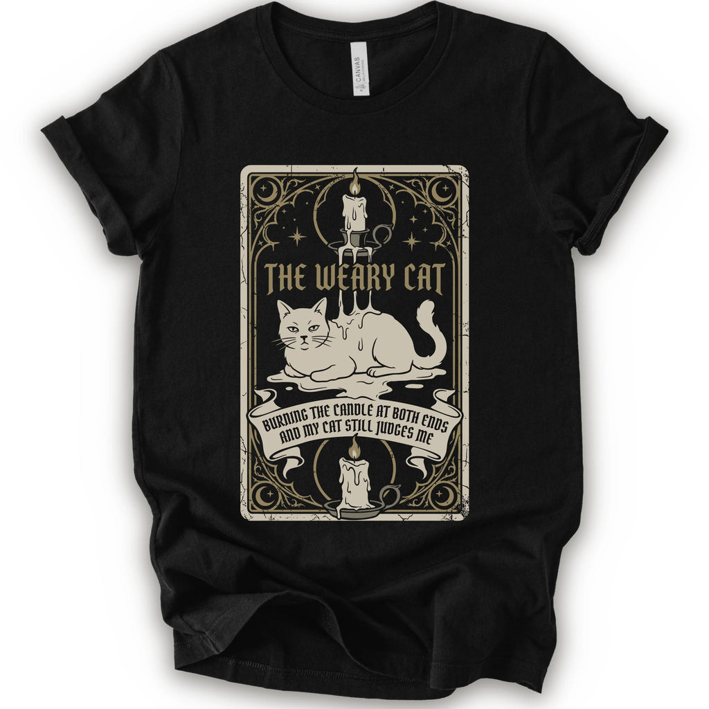 A black short-sleeve crewneck t-shirt with a vintage-style graphic print on the front. The print features a white cat lying down inside a decorative frame with candles, stars, and the text 'THE WEARY CAT' and 'BURNING THE CANDLE AT BOTH ENDS AND MY CAT STILL JUDGES ME.