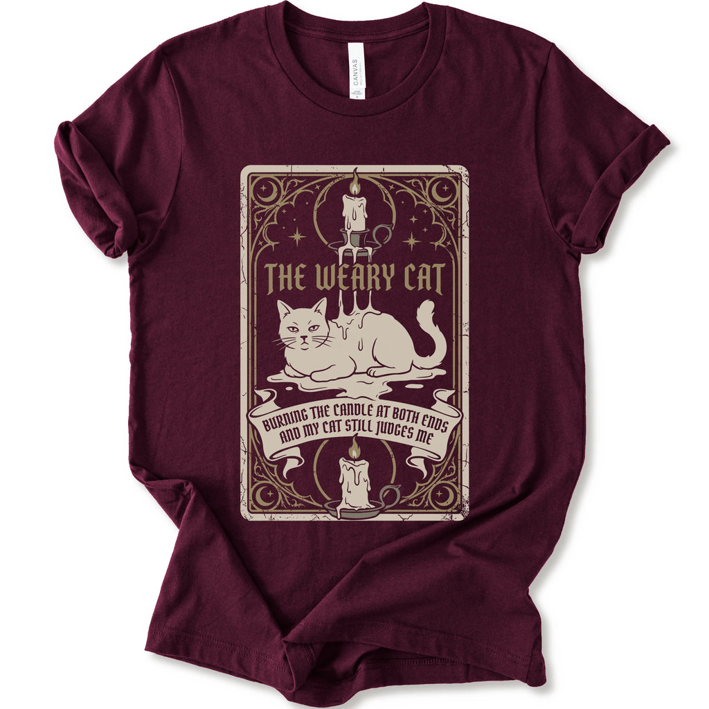 A maroon short-sleeve crewneck t-shirt with a vintage-style graphic print on the front. The print features a white cat lying down inside a decorative frame with candles, stars, and the text 'THE WEARY CAT' and 'BURNING THE CANDLE AT BOTH ENDS AND MY CAT STILL JUDGES ME.