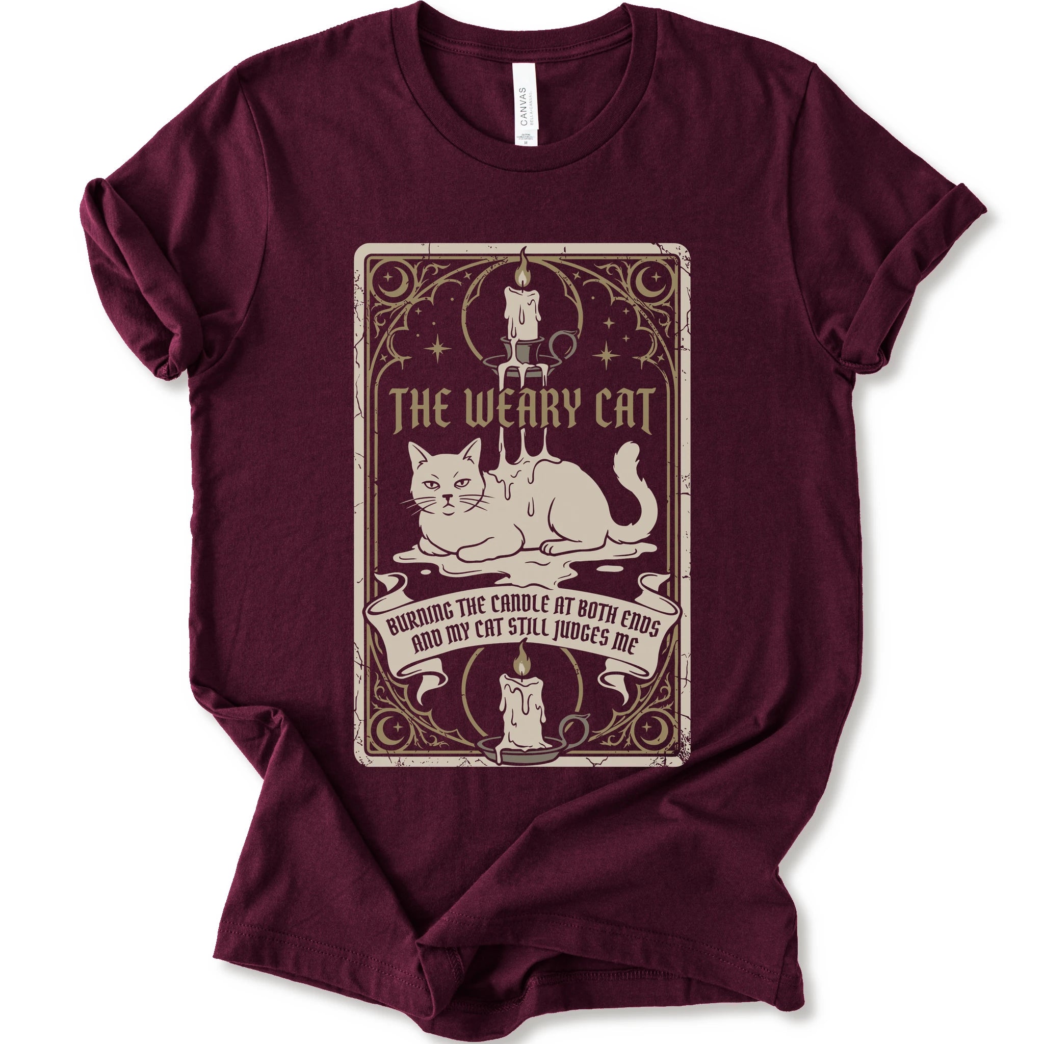 A maroon short-sleeve crewneck t-shirt with a vintage-style graphic print on the front. The print features a white cat lying down inside a decorative frame with candles, stars, and the text 'THE WEARY CAT' and 'BURNING THE CANDLE AT BOTH ENDS AND MY CAT STILL JUDGES ME.