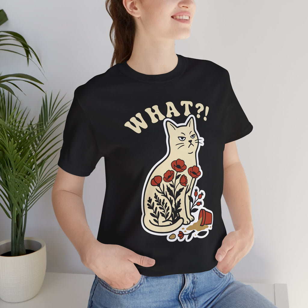 A woman wearing a black graphic t-shirt with a cartoon cat and the text 'WHAT?!' printed on the front, paired with blue jeans, standing next to a potted plant.