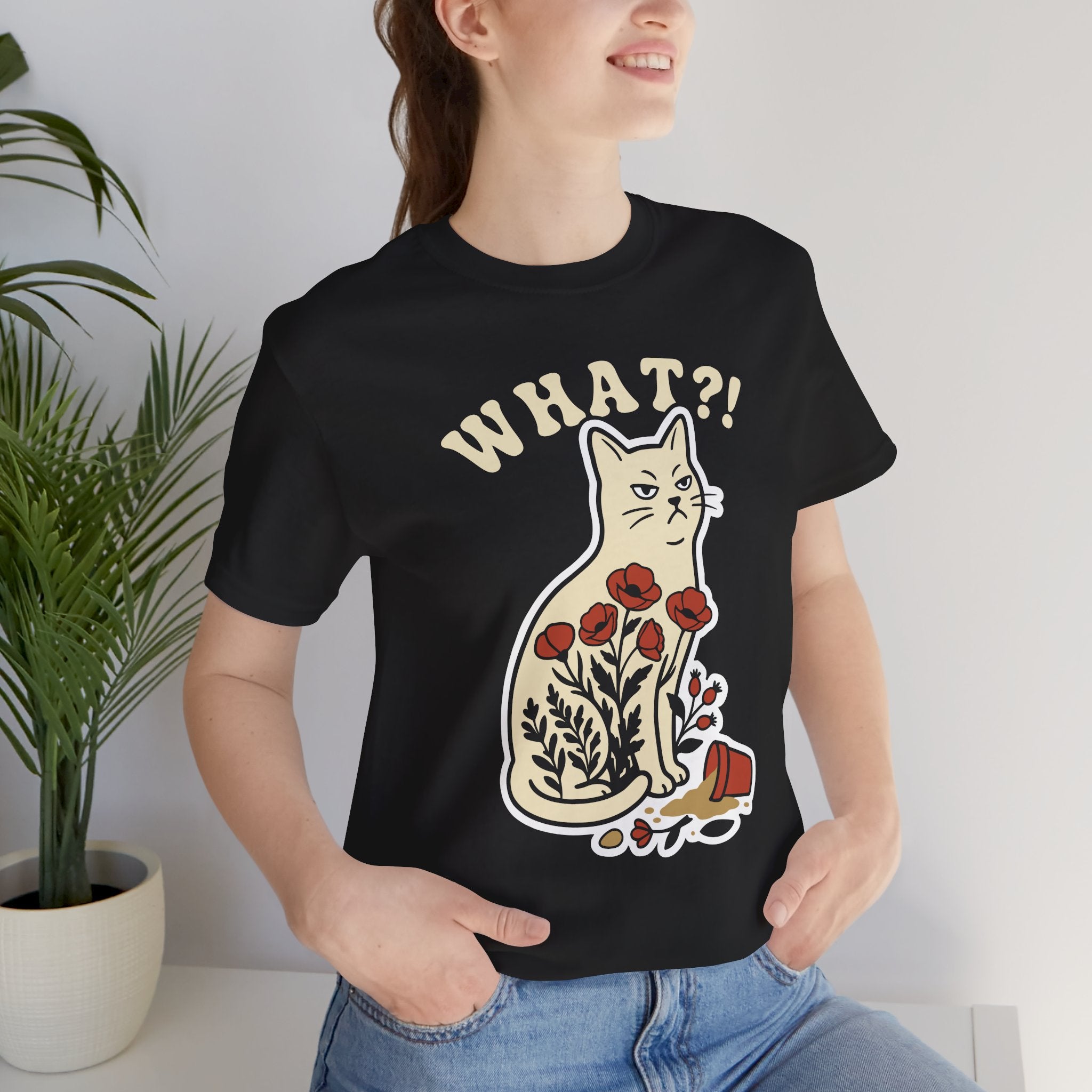 A woman wearing a black graphic t-shirt with a cartoon cat and the text 'WHAT?!' printed on the front, paired with blue jeans, standing next to a potted plant.