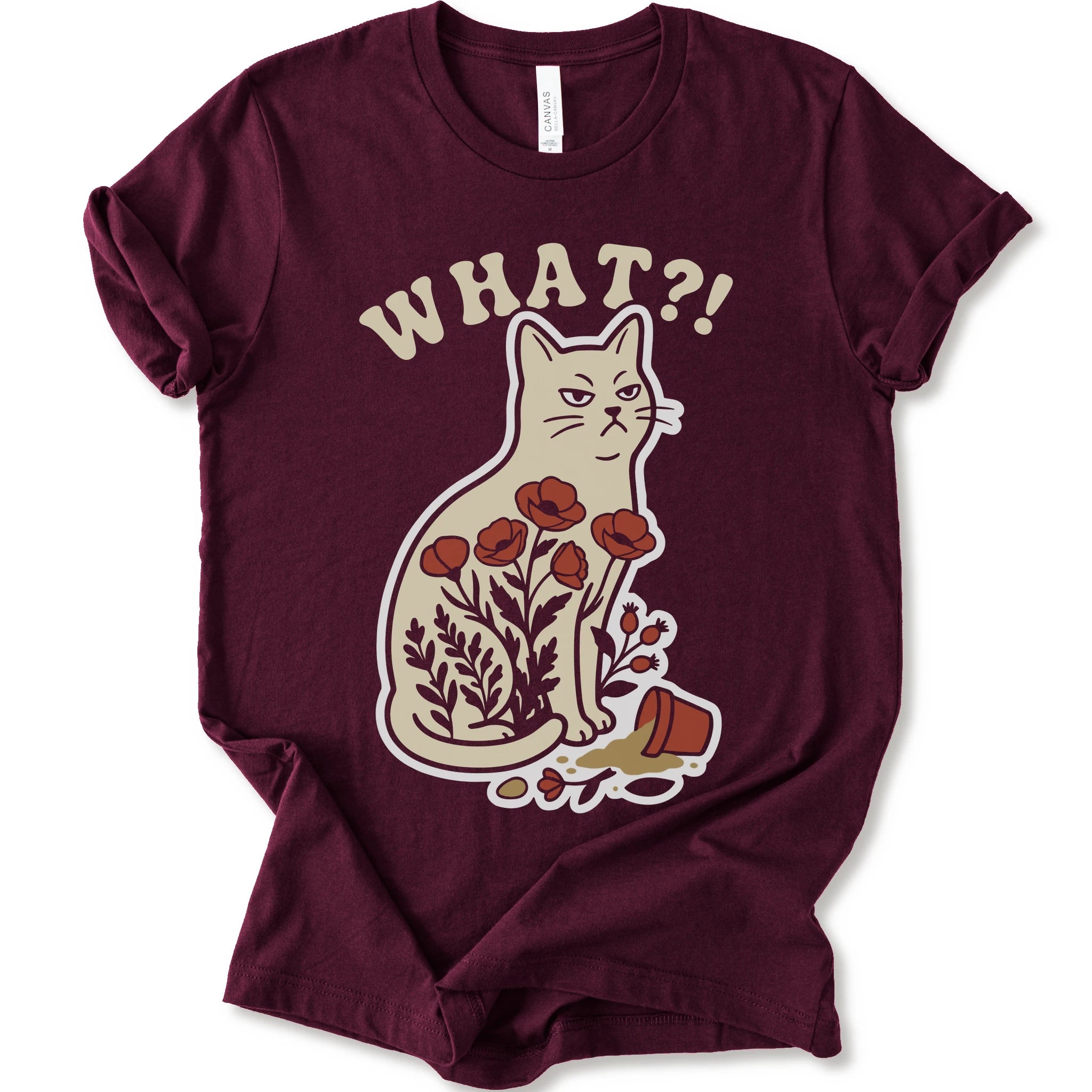 Maroon graphic t-shirt with “WHAT?!” text above an unimpressed cream cat with red flowers beside a knocked-over plant pot